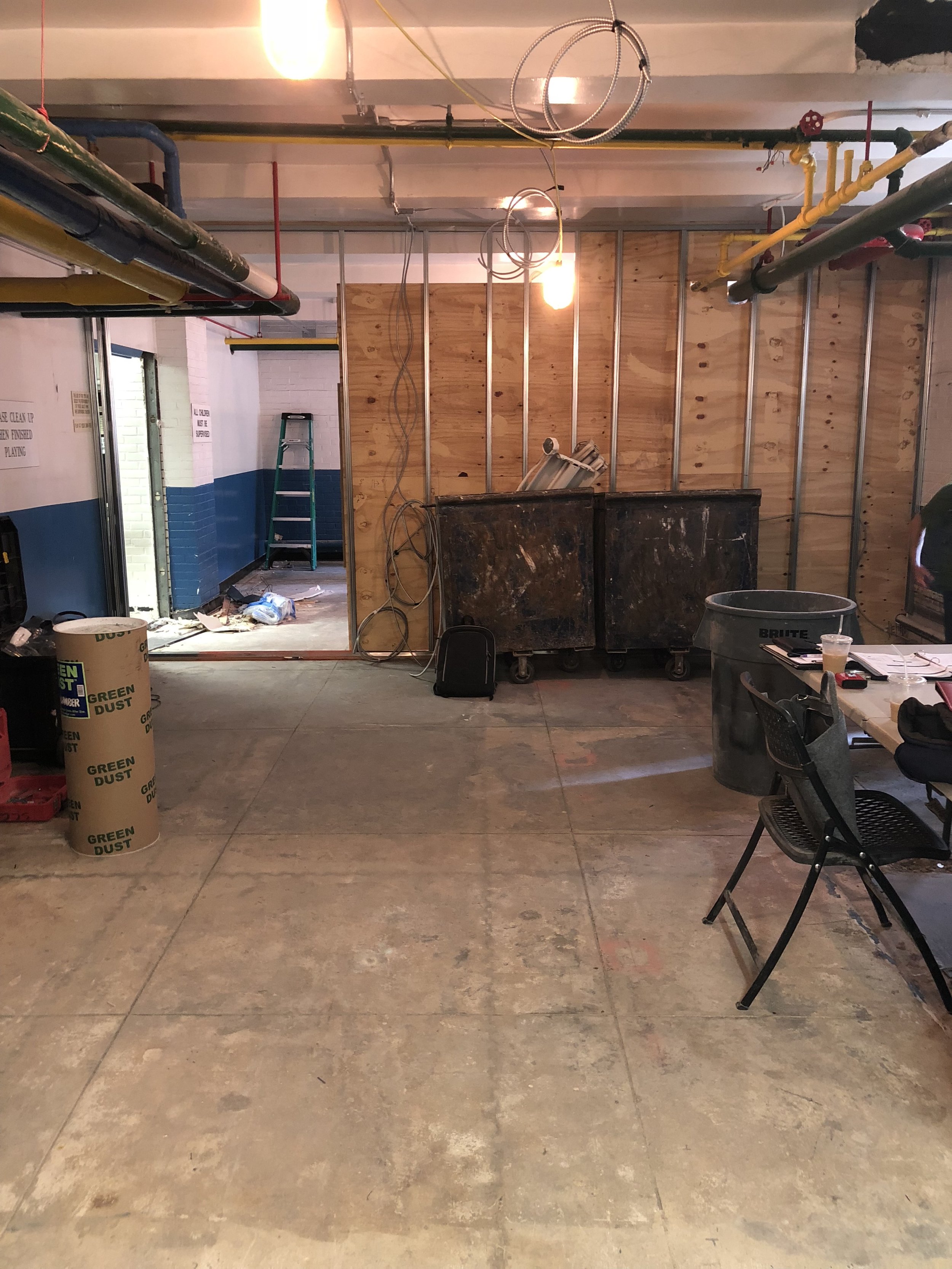 Interior of a space under construction or renovation, with exposed wooden wall framing, electrical wiring, and piping on the ceiling. There are construction materials including a large trash bin, a green dust barrel, a ladder, and a desk with chairs and office supplies.