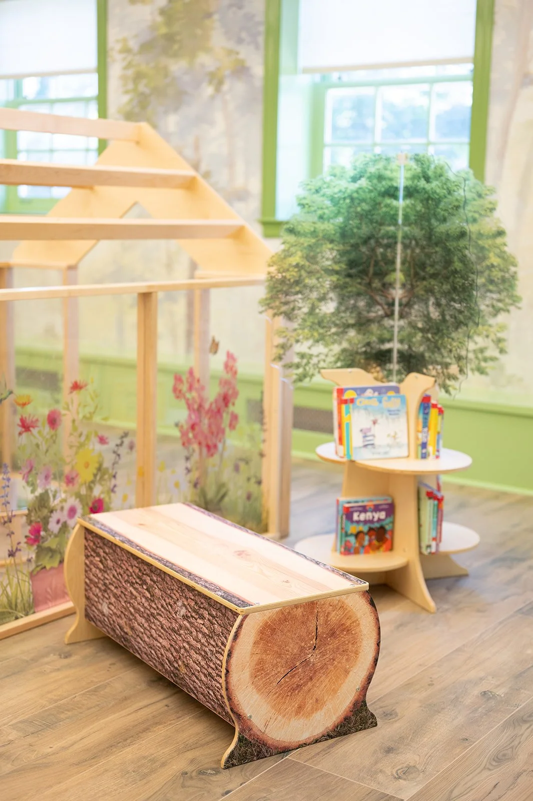 A children's reading nook with a tree-shaped bookshelf, a wooden log bench, and a backdrop of greenery and flowers on a glass window.