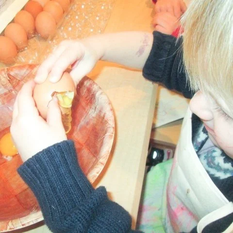 Child cracking an egg into a bowl on a kitchen counter surrounded by eggs and cooking tools.