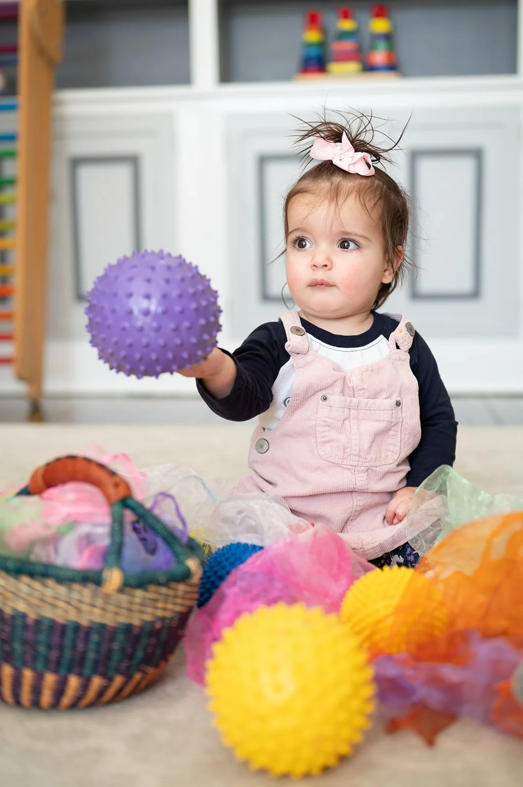 A young girl with a pink bow in her hair, wearing a black long-sleeve shirt and pink overalls, sitting on the floor surrounded by colorful spiky sensory balls and a small woven basket, holding out a purple ball.
