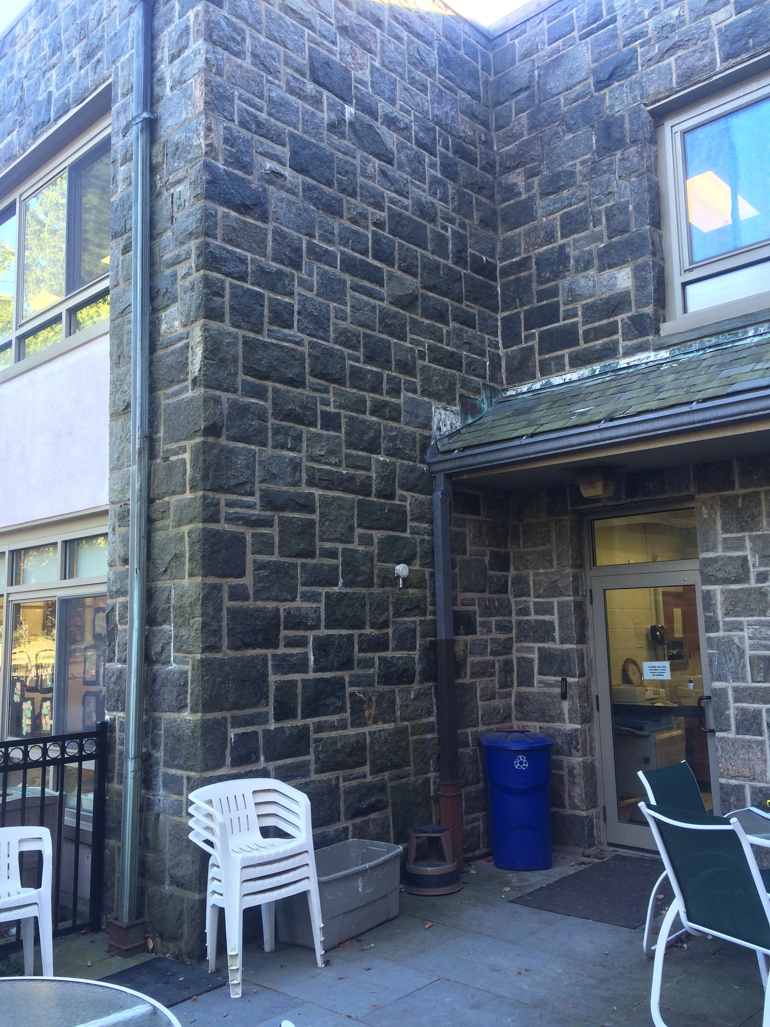 Exterior view of a building with stone walls, a glass door, outdoor seating, a blue recycling bin, and windows.