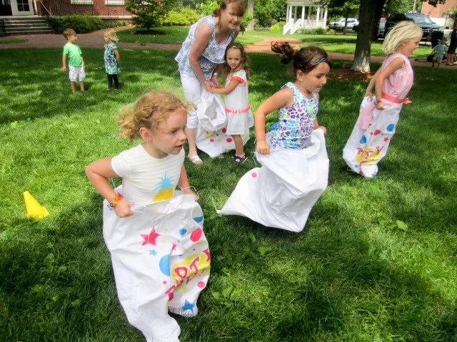 Children playing sack race on a grassy field during a celebration, with a woman supervising.