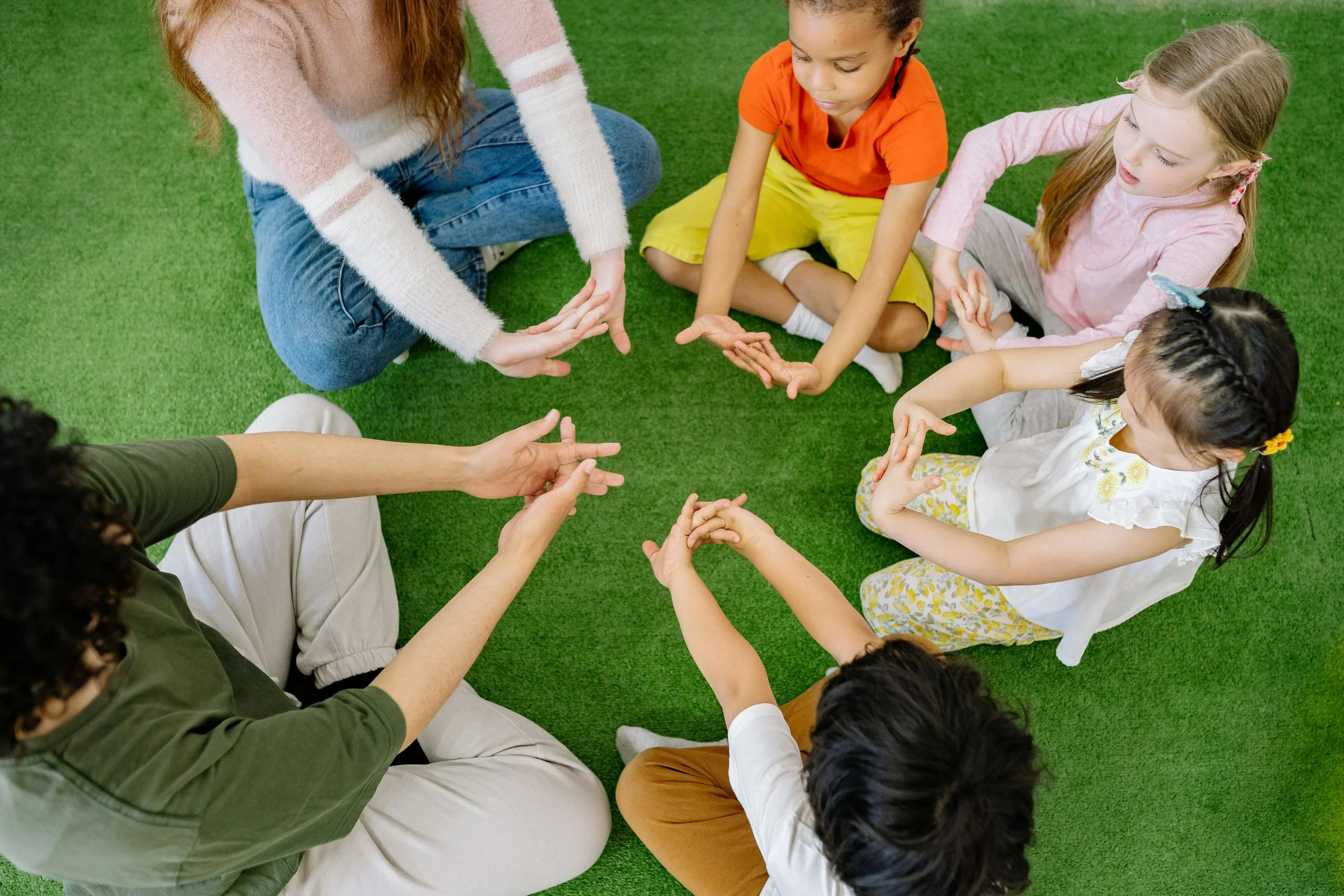 Group of children and a woman sitting on green artificial grass, engaging in a circle activity with their hands.