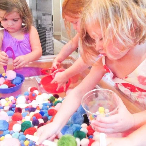 Children playing with colorful pom-poms and craft supplies at a table.