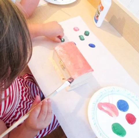 Child painting a wooden house with pink paint, surrounded by paint bottles and palette with red, green, and blue colors.