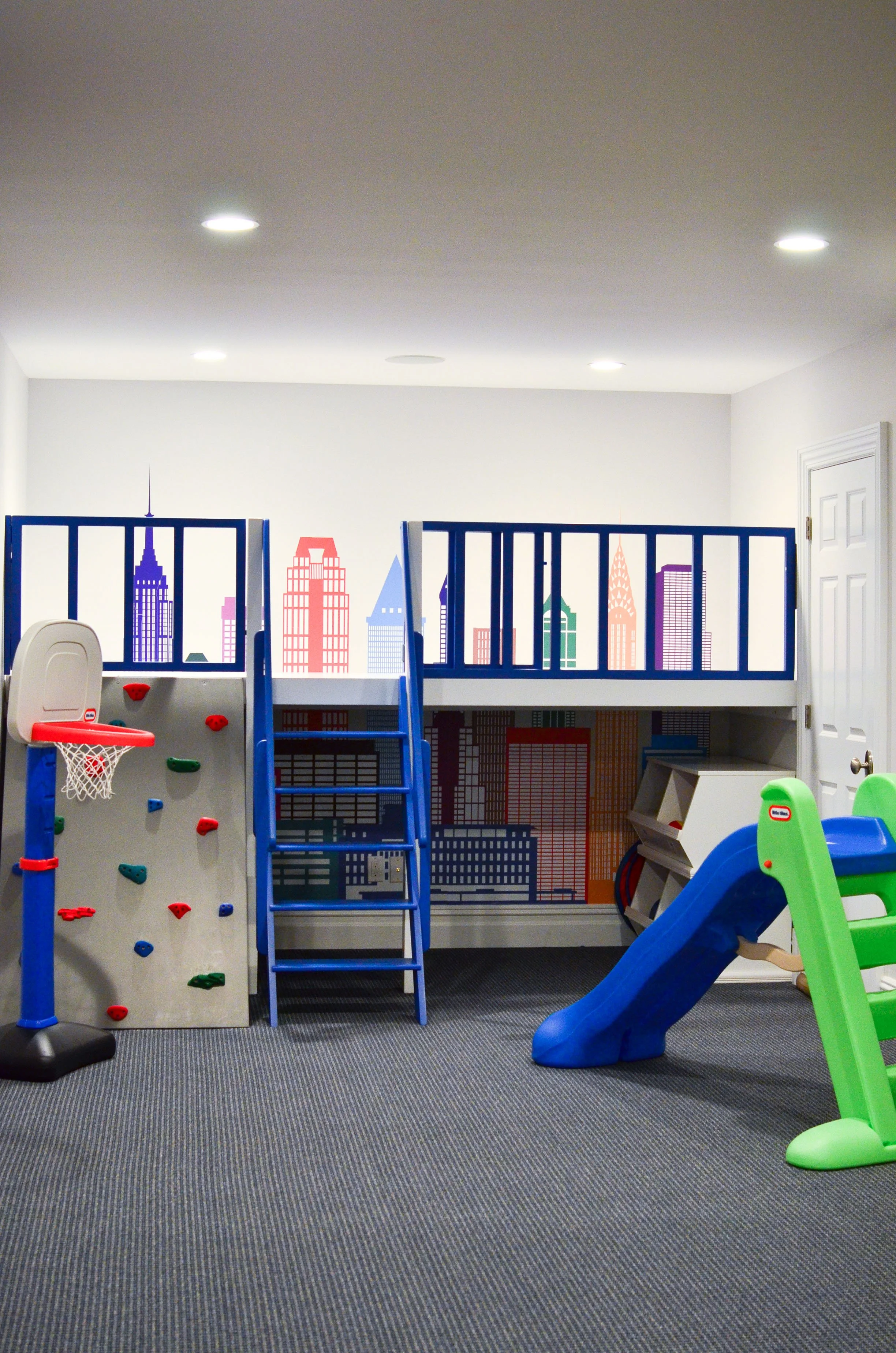 Indoor playroom with a blue loft, multicolored climbing wall, small blue slide, and cityscape wall mural.