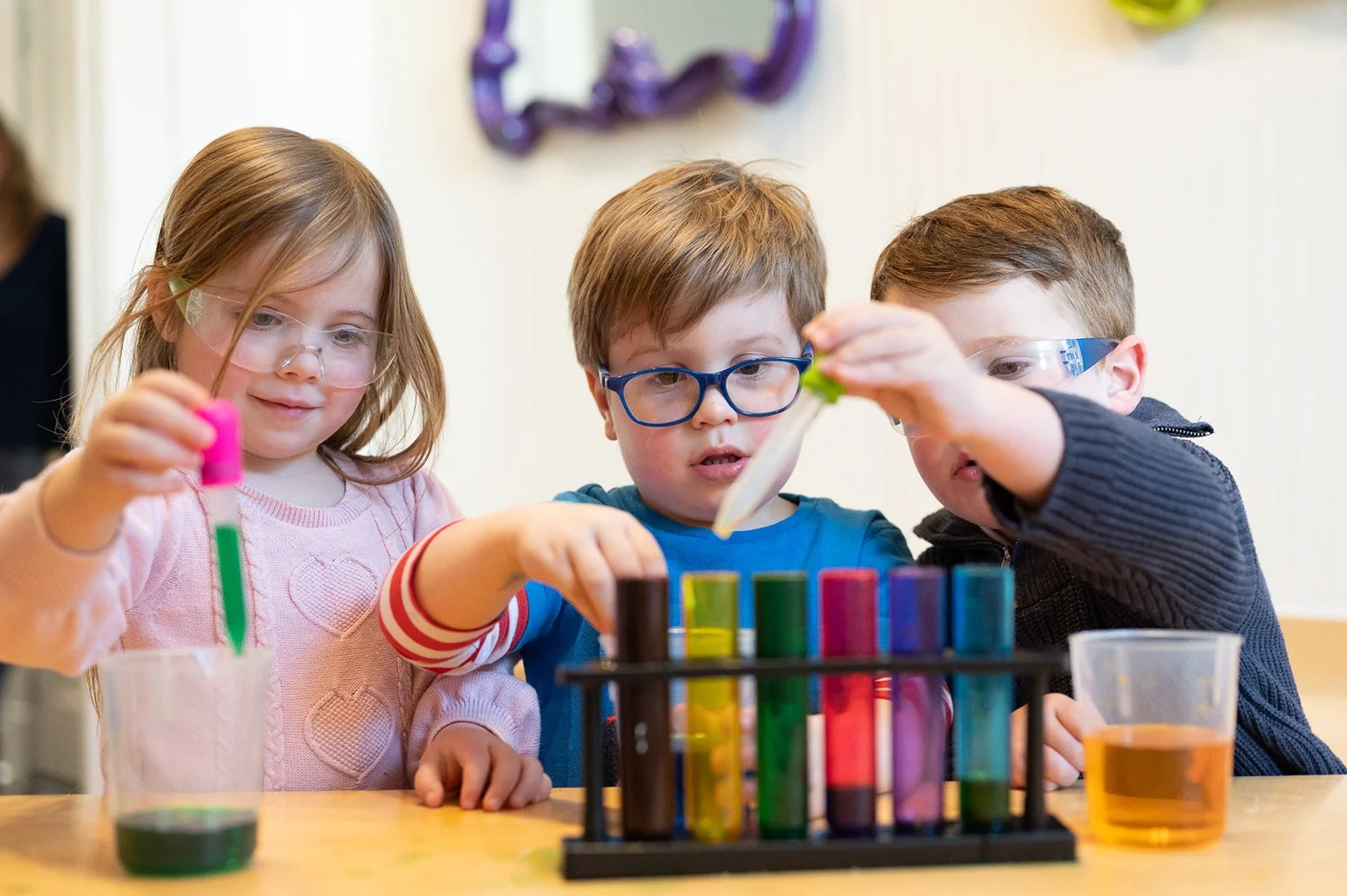 Three children wearing safety goggles conducting a science experiment with test tubes and liquids at a table.