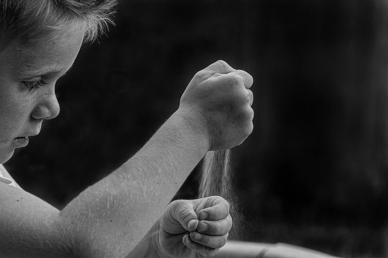 A black and white photo of a young girl with short hair, seen from the side, squeezing a handful of sand or dust with her right hand.