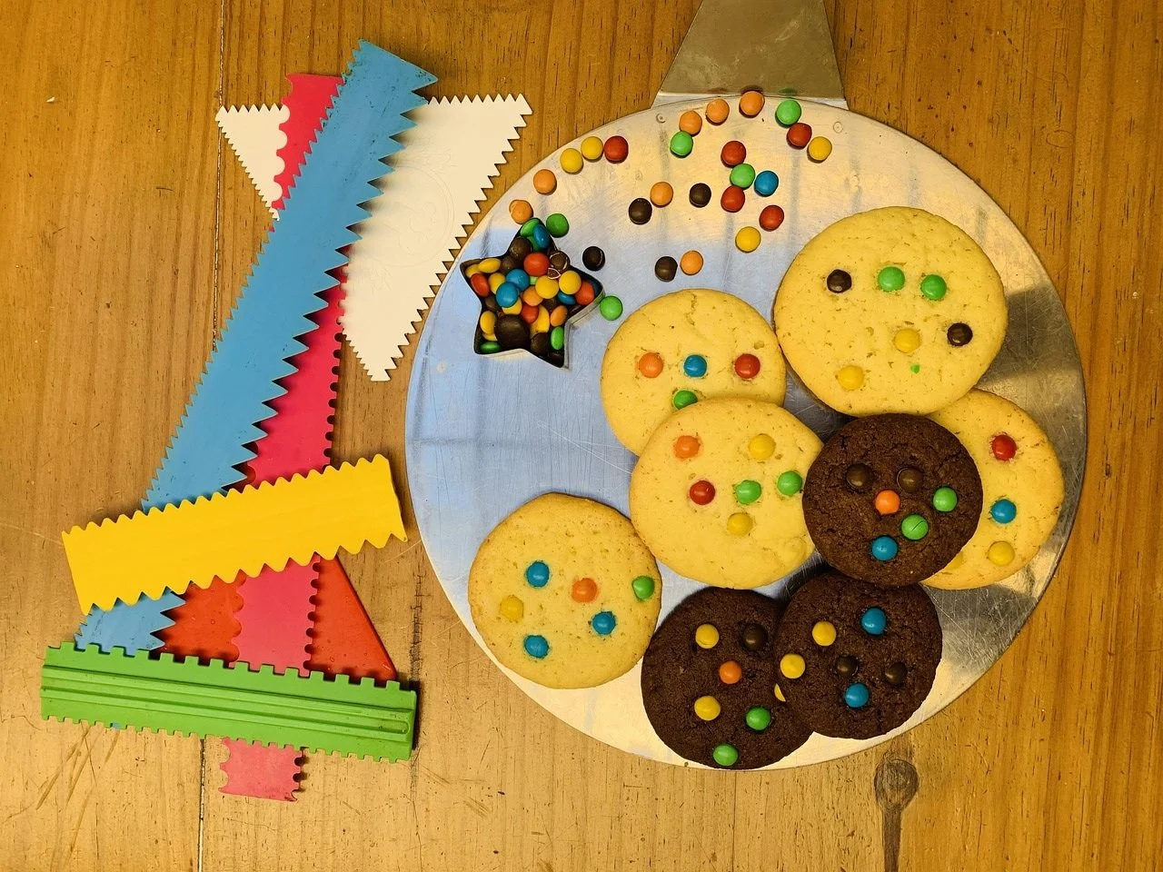 Colorful sprinkles on vanilla and chocolate cookies on a round metal tray, with some sprinkles spilled on the tray and various colorful zigzag-edged paper strips on a wooden surface nearby.