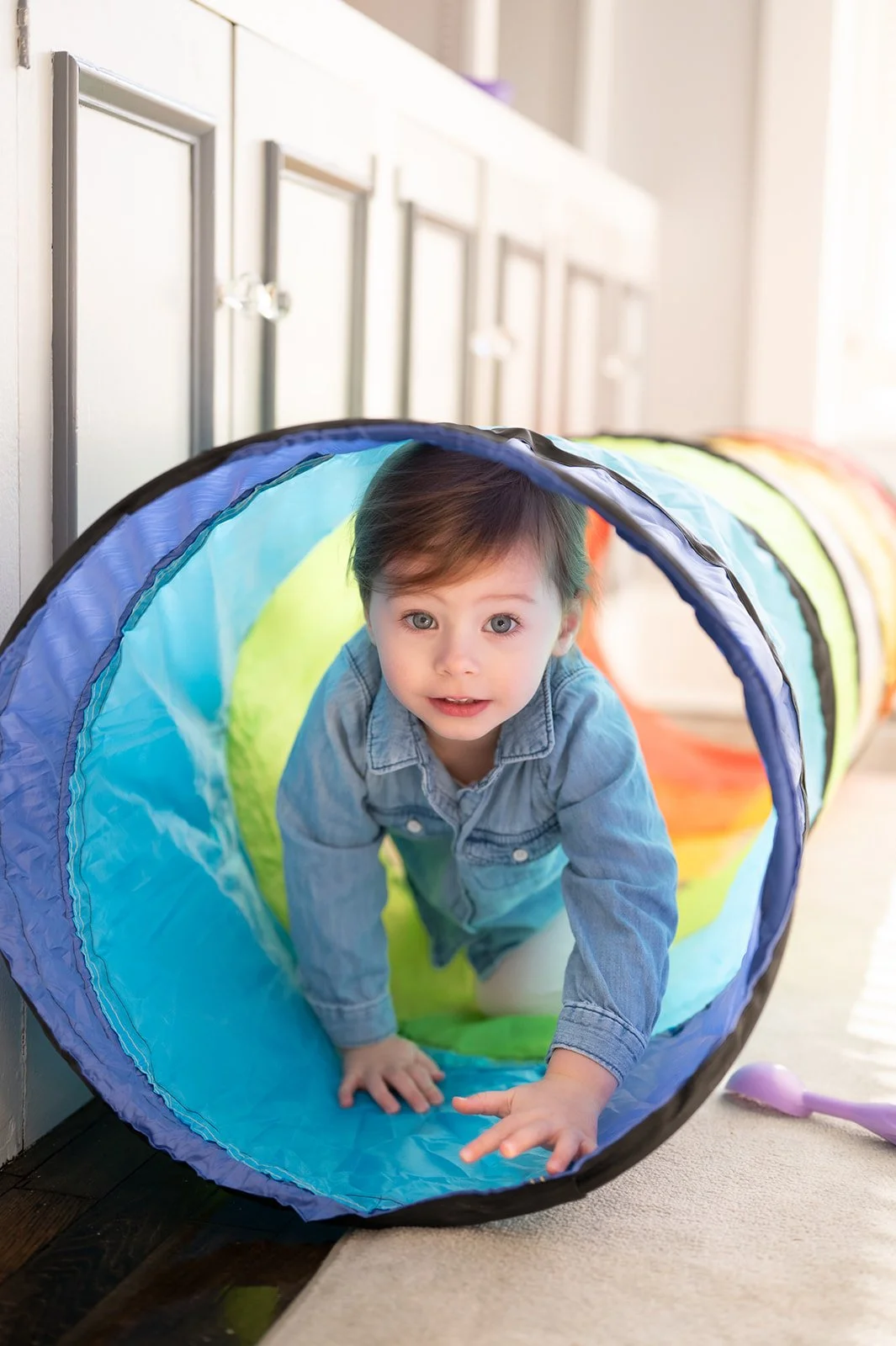 A young child crawling through a colorful fabric tunnel on a carpeted floor in a sunny room.