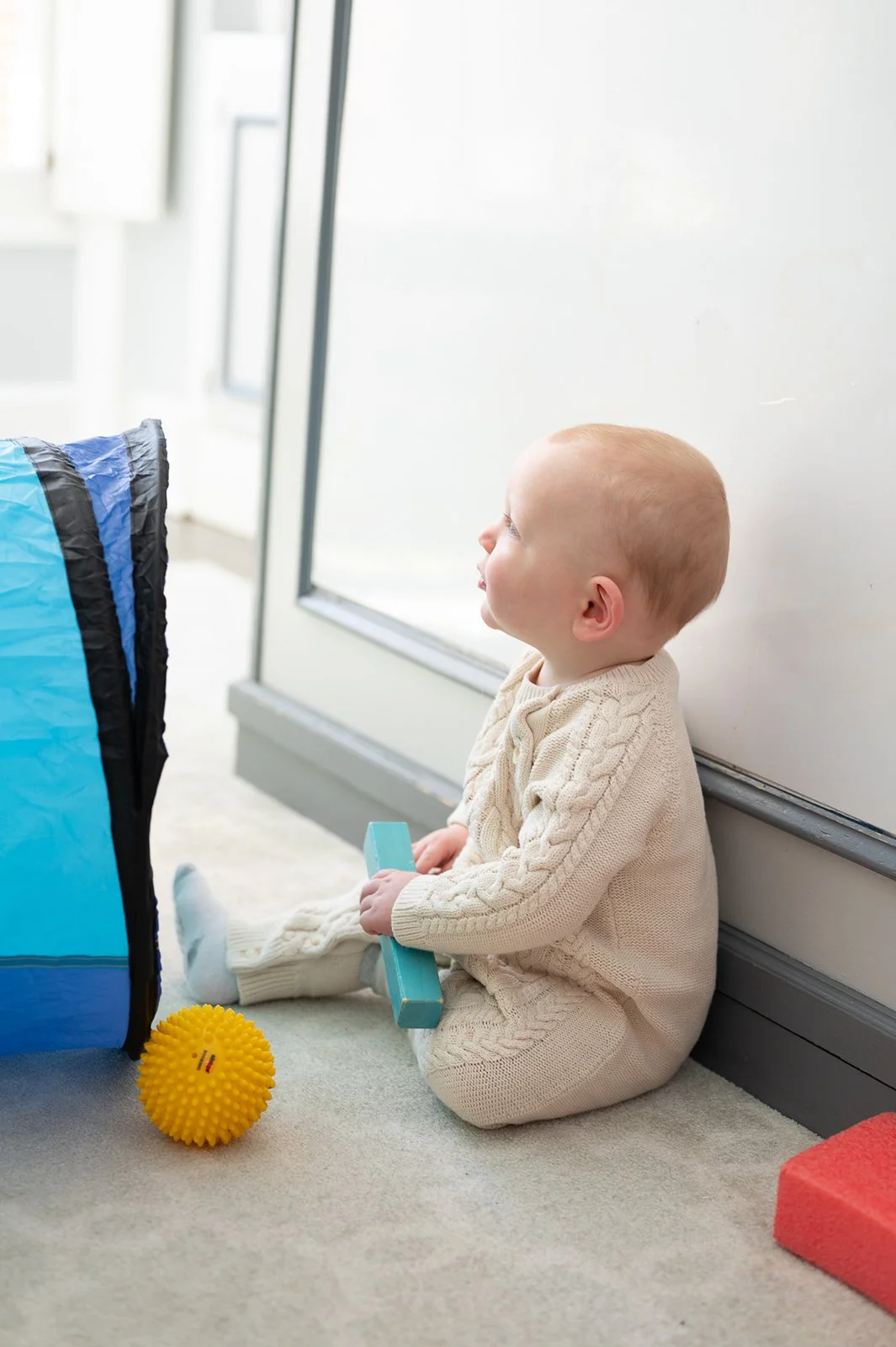 A young child with a bald head, sitting on a carpeted floor and looking to the left, holding a blue toy. The child is wearing a beige knitted sweater and matching pants. There is a yellow textured ball in front of them and foam blocks nearby, with a large blue and black tent structure on the left. The background includes a white wall and a window letting in natural light.
