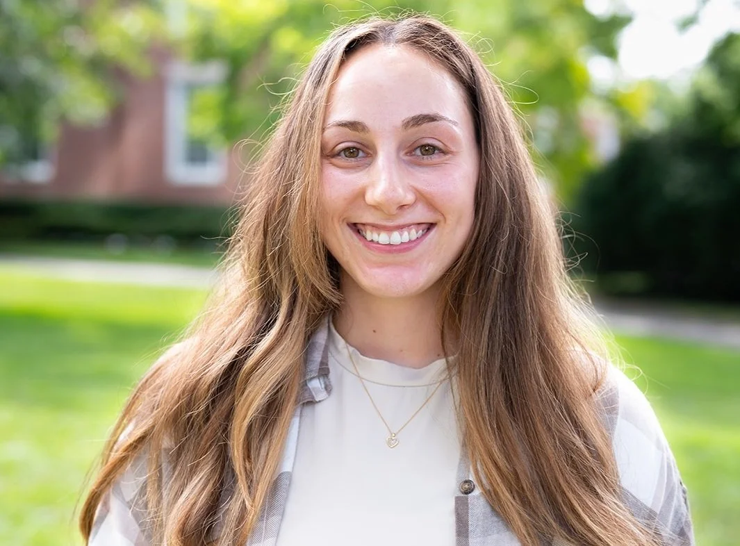 A young woman with long brown hair smiling outdoors in a park with green trees and grass in the background.