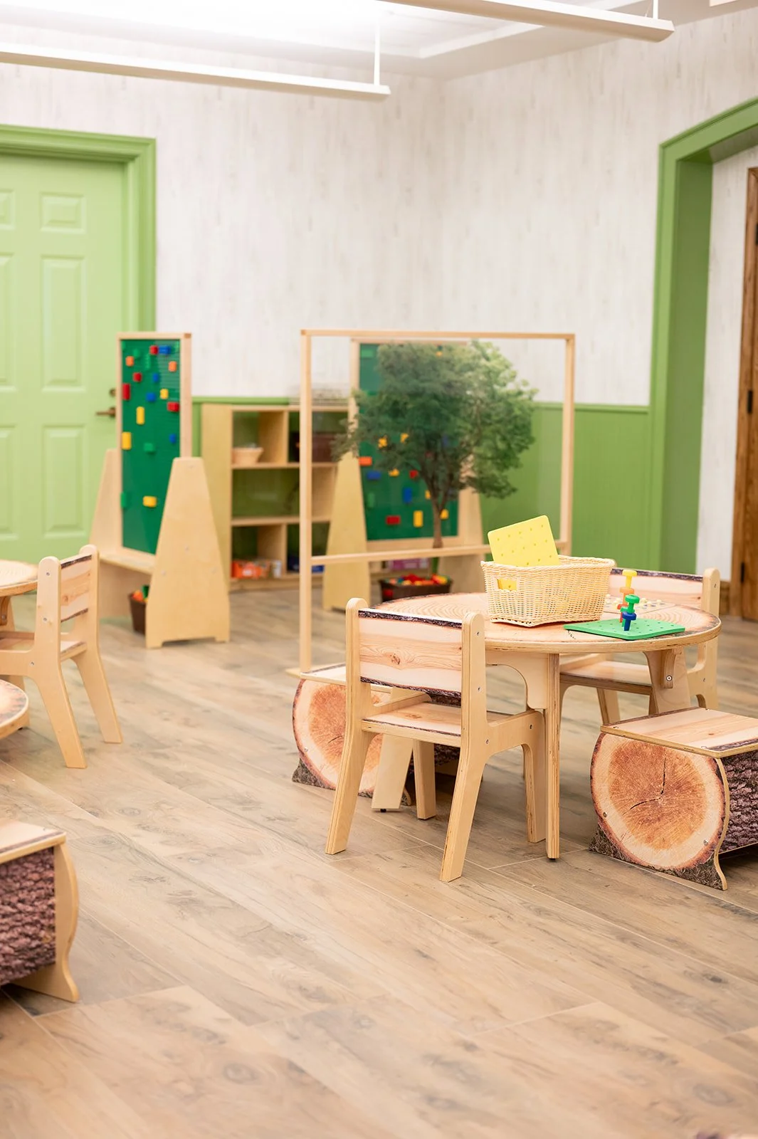A children's classroom decorated with natural wood furniture, a small round table with three chairs, and a log-shaped bench. There are indoor play structures with a climbing wall and a tree prop, with a green door and light-colored walls in the background.