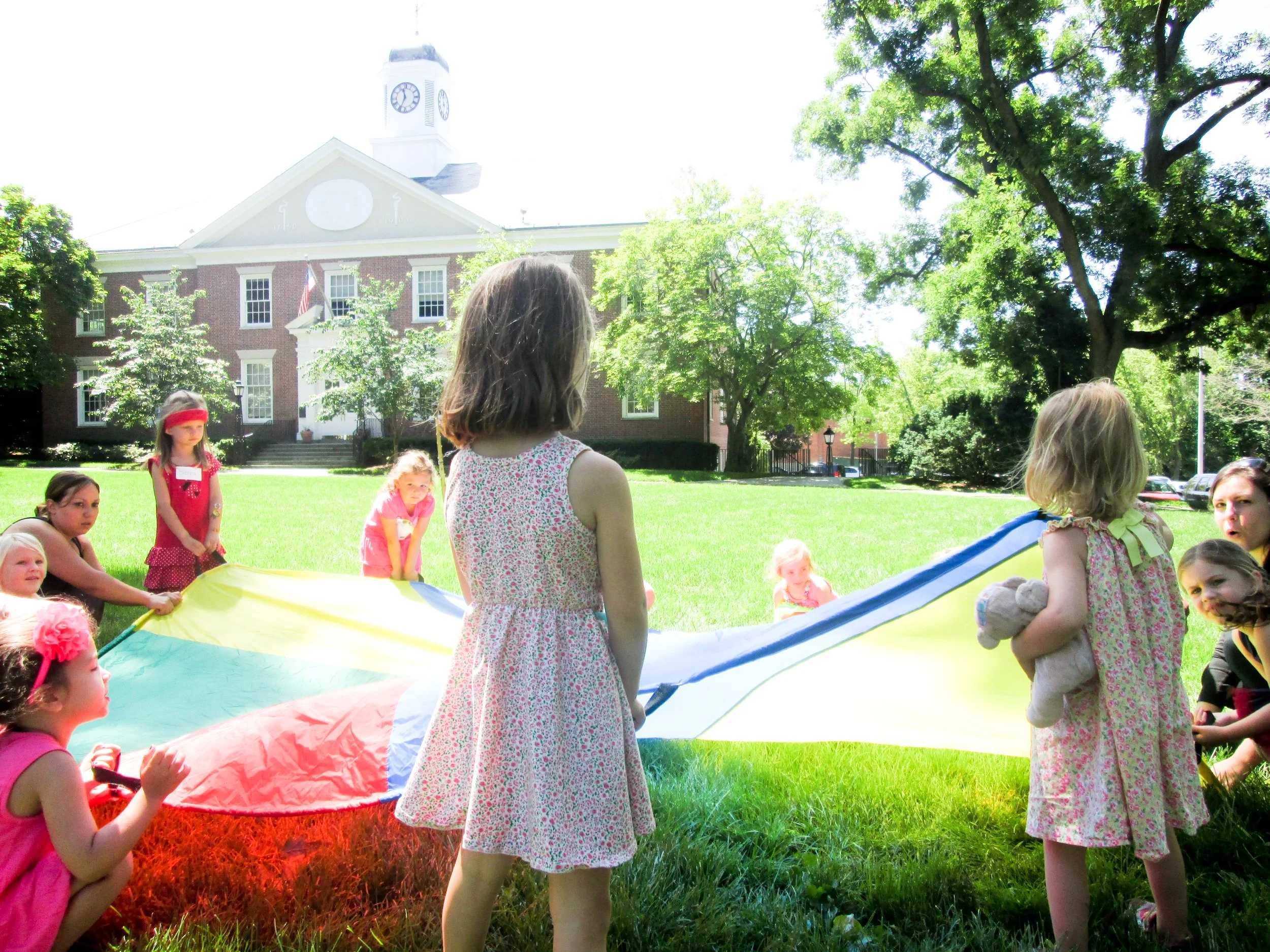 Group of children playing with a colorful parachute outside on a grassy lawn with a large brick building and tall trees in the background.
