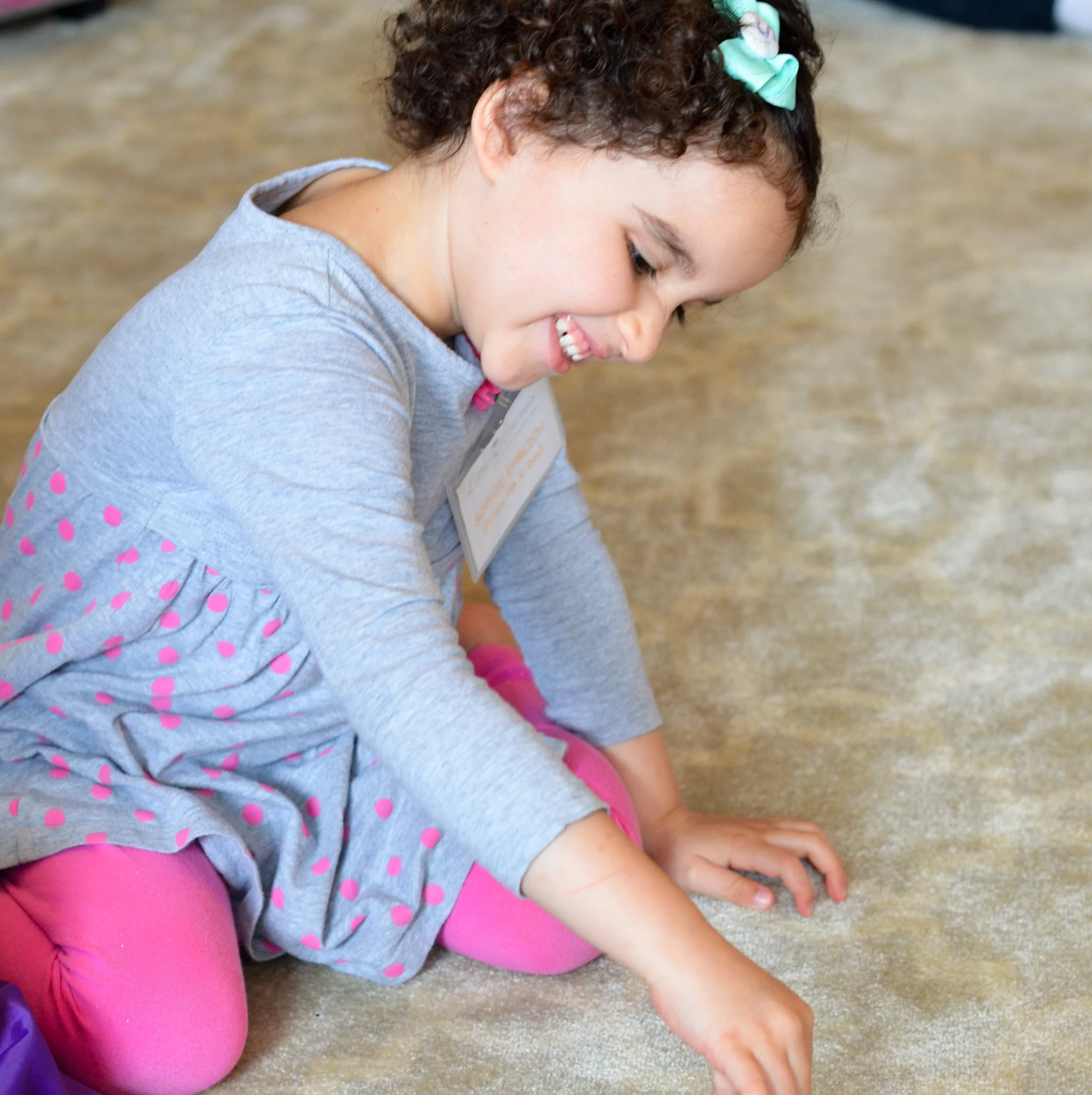 A young girl with curly hair, wearing a gray dress with pink polka dots and pink tights, sitting cross-legged on the floor, smiling and reaching towards the ground.