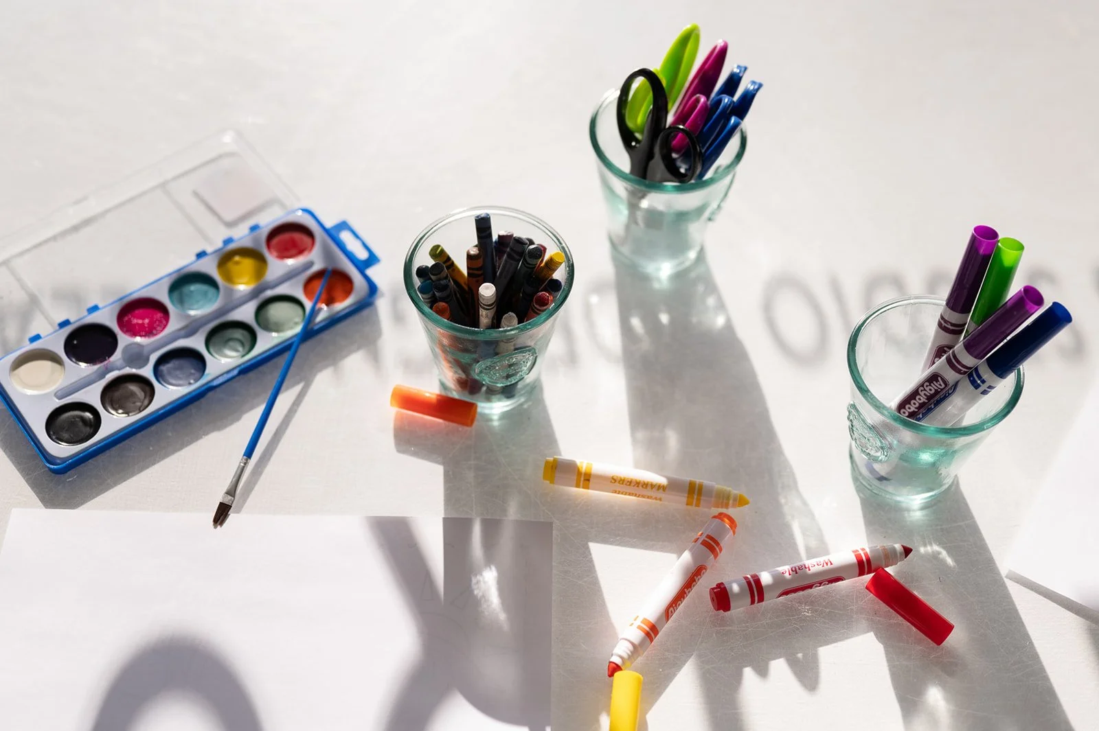 Assorted arts and crafts supplies on a white surface, including watercolor paints, paintbrush, colored markers in clear glasses, and crayon pieces.