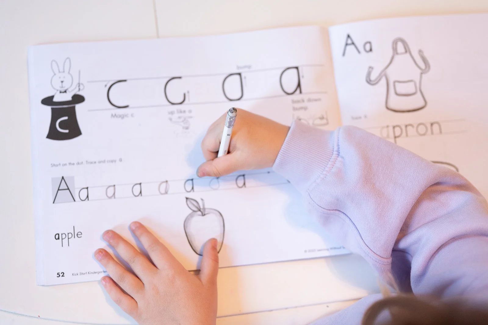 A child is tracing and coloring a worksheet with alphabet and word exercises, including a drawing of an apple.