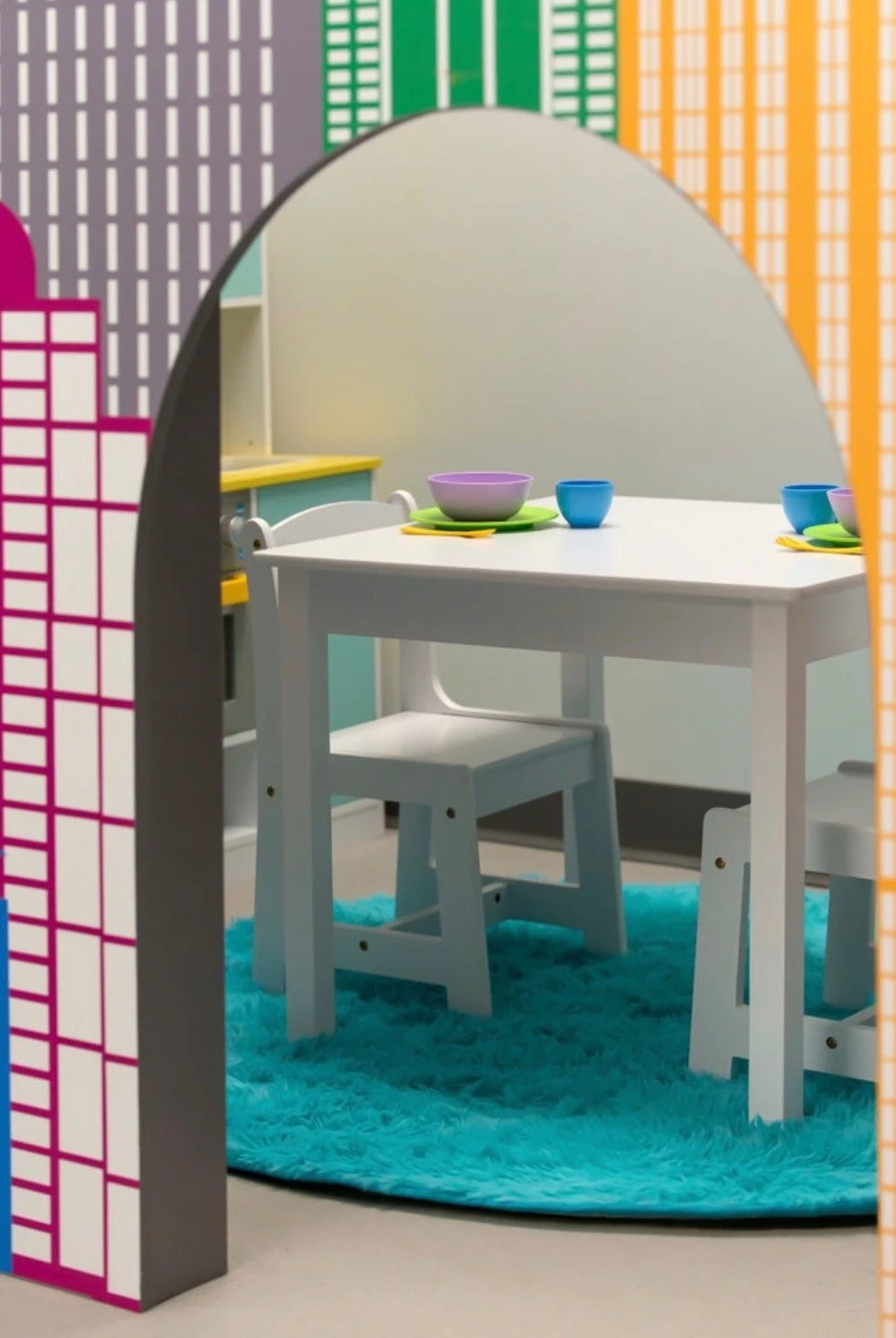A children's play area with a rectangular white table, two small white chairs, colorful tableware including bowls and cups, a bright blue shaggy rug, and a cityscape mural with multicolored buildings on the wall.