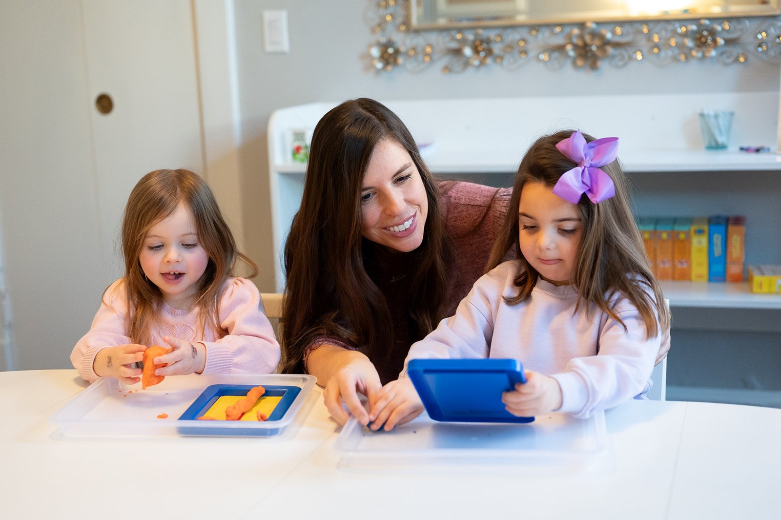 A woman and two young girls sitting at a table, engaging with a tablet and playdough. The girl on the left has long red hair and is holding orange playdough, while the girl on the right has brown hair with a purple bow and is looking at the tablet.