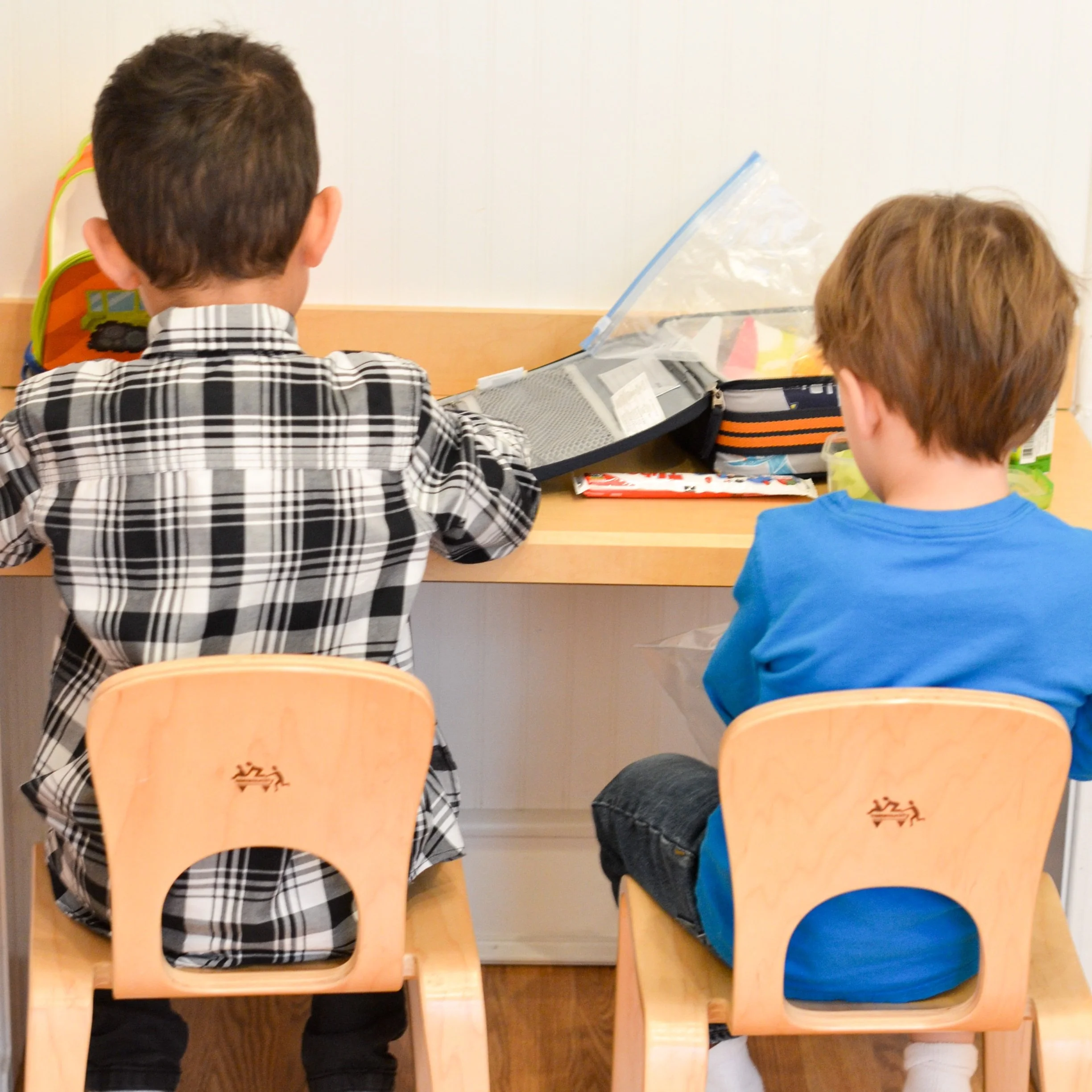 Two children sitting at a desk viewed from behind, with school supplies on the desk and empty chairs.