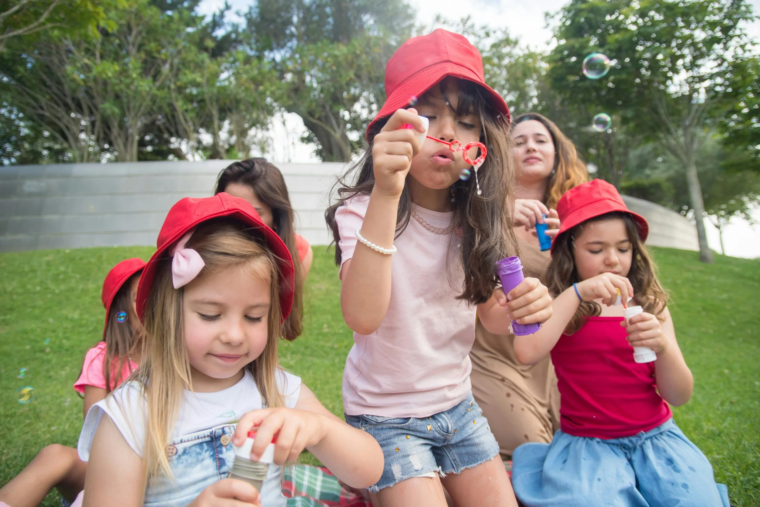 Children and an adult girl blowing bubbles outdoors in a park, wearing red hats and colorful clothing.