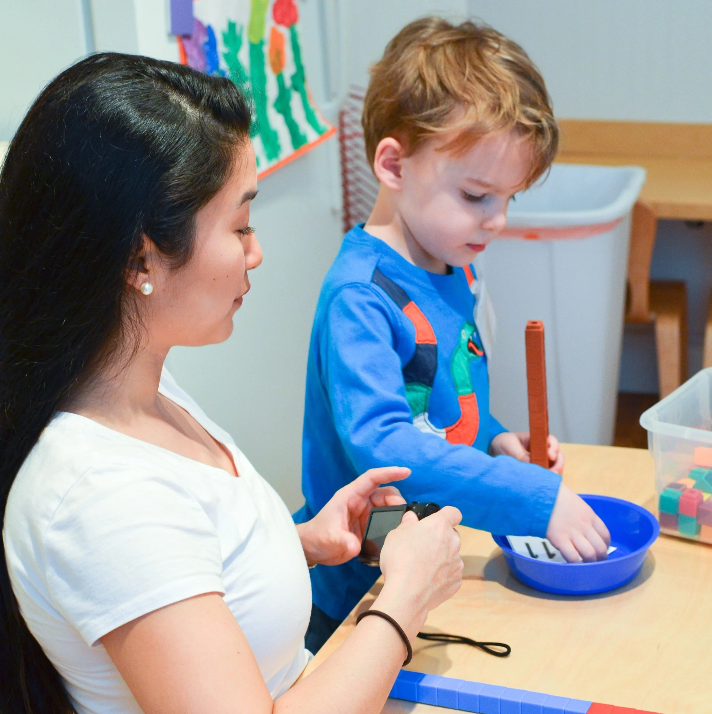 A woman and a young boy engaging in a hands-on activity at a table in a classroom. The woman is holding a smartphone, and the boy is focused on a small object in a blue bowl, with colorful building blocks nearby.