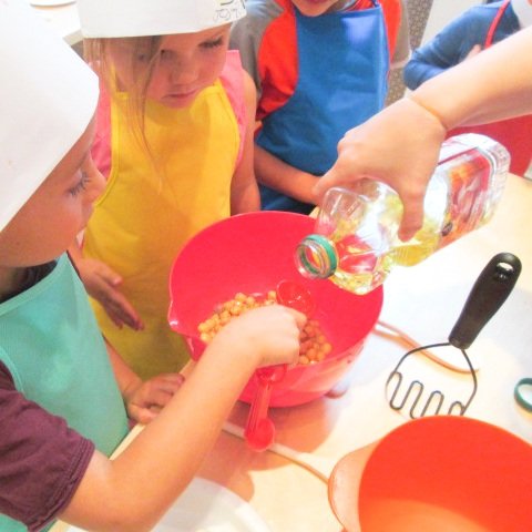 Children wearing aprons and chef hats watching an adult pouring a bottle of oil into a mixing bowl with popcorn, in a kitchen setting.