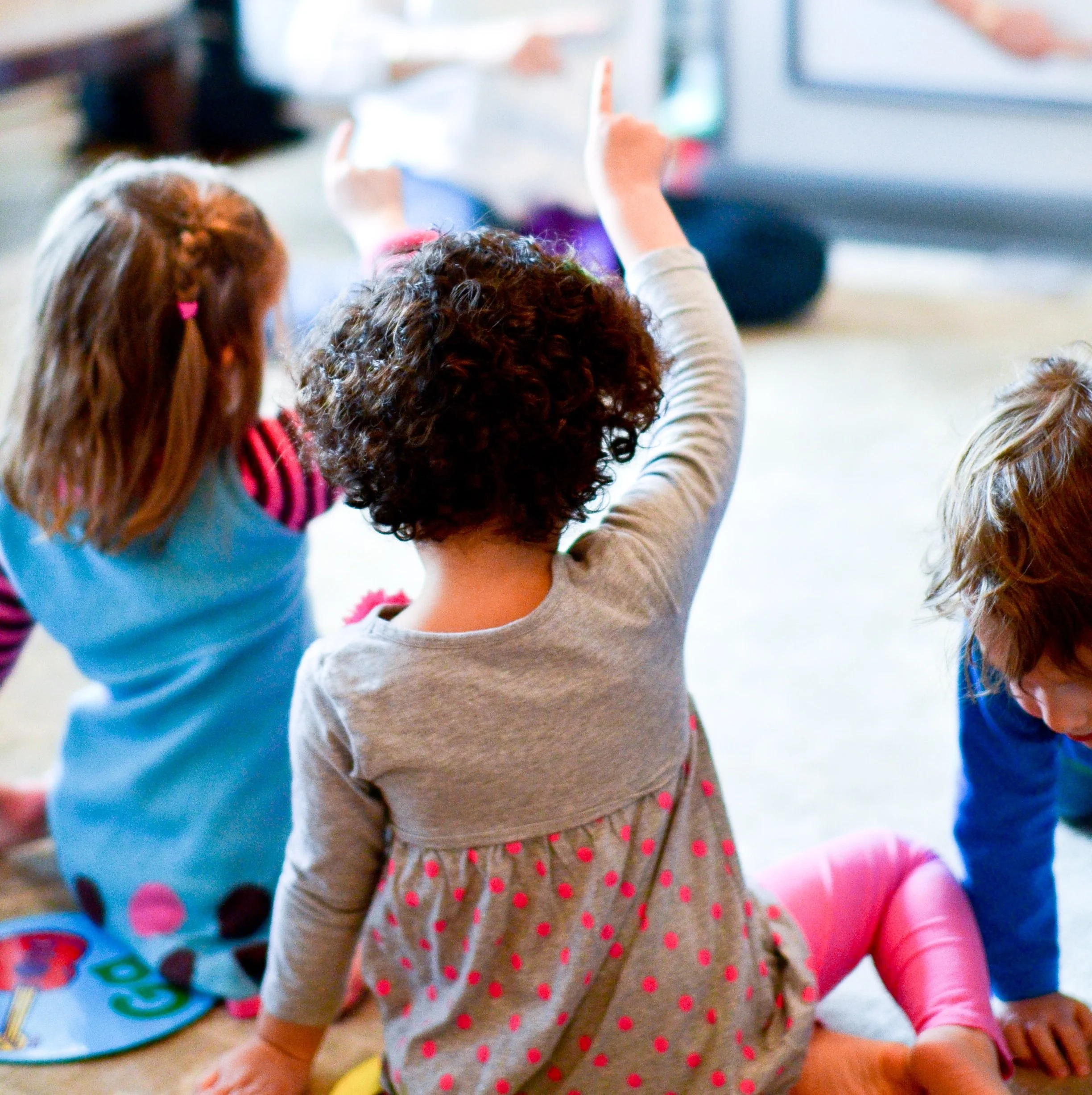 Three children sitting on the floor watching a television, one girl with curly hair and a gray dress with pink dots raising her hand.