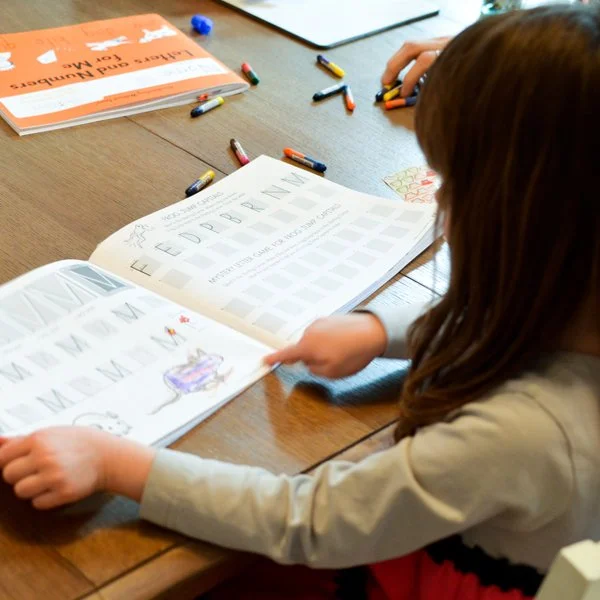 A young girl is practicing the alphabet in a workbook at a wooden table, with scattered crayons and a coloring book nearby.