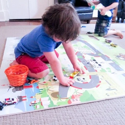 Child playing with toy cars on a colorful play mat while kneeling on the carpet, with a basket nearby.