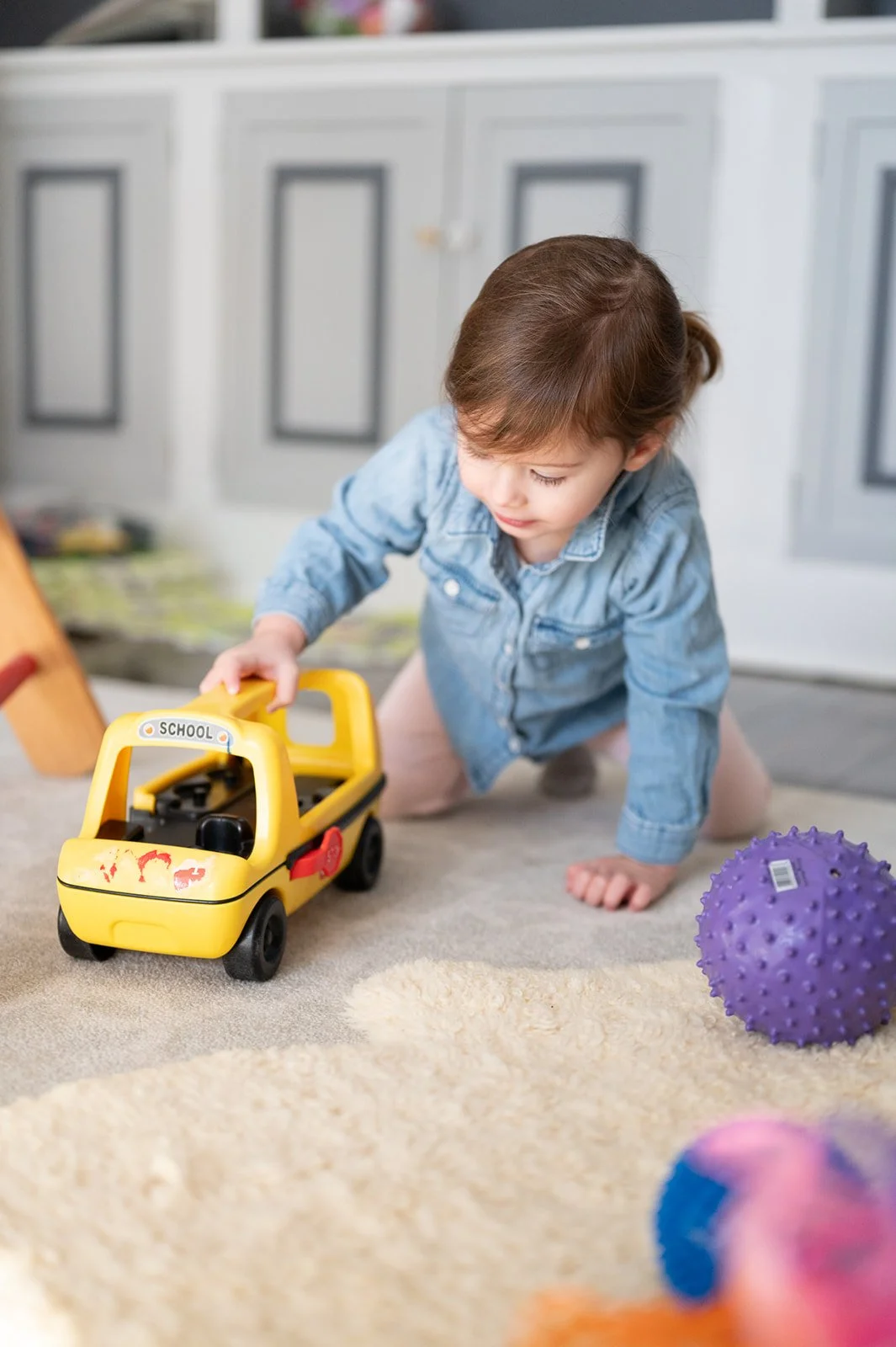 A young child playing with a yellow toy school bus on a beige carpeted floor in a room with light gray cabinets in the background. There is a purple spiky ball and some other toys visible on the floor.