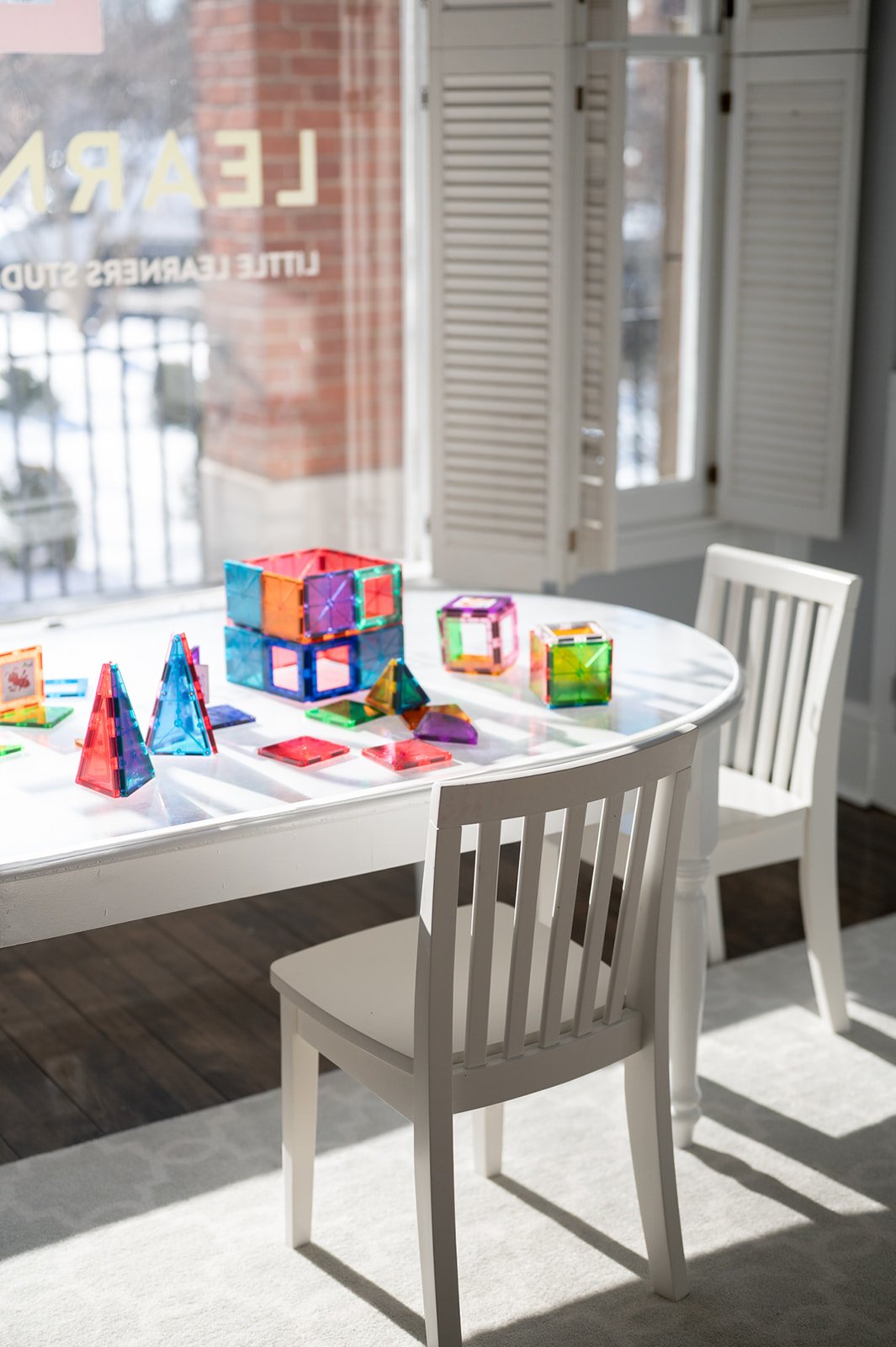 Colorful magnetic building tiles arranged on a white dining table with open window shutters in the background.