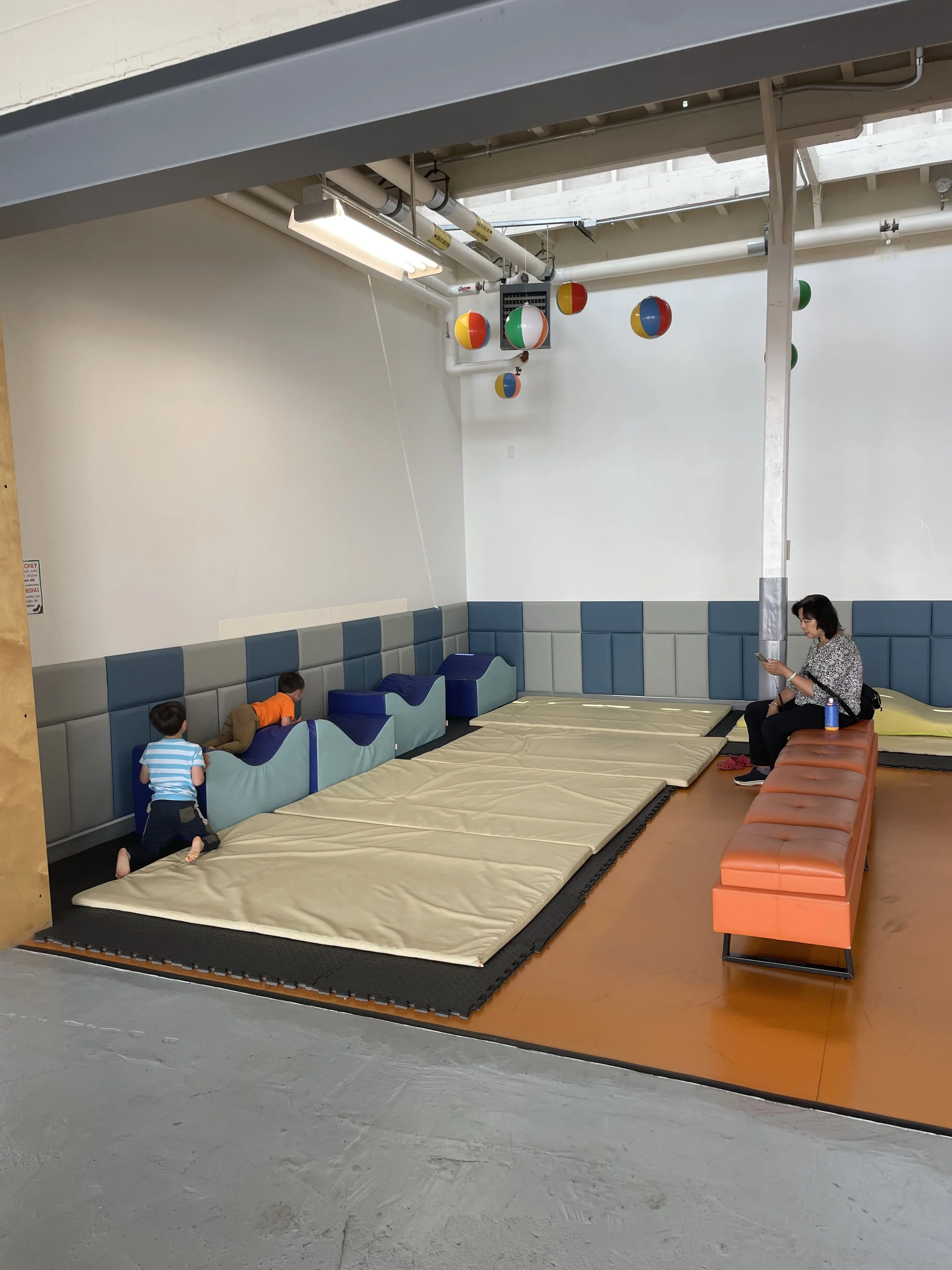 Indoor play area with mats, padded walls, and colorful beach balls hanging from the ceiling. Two children are playing, and a woman is sitting on an orange bench looking at her phone.