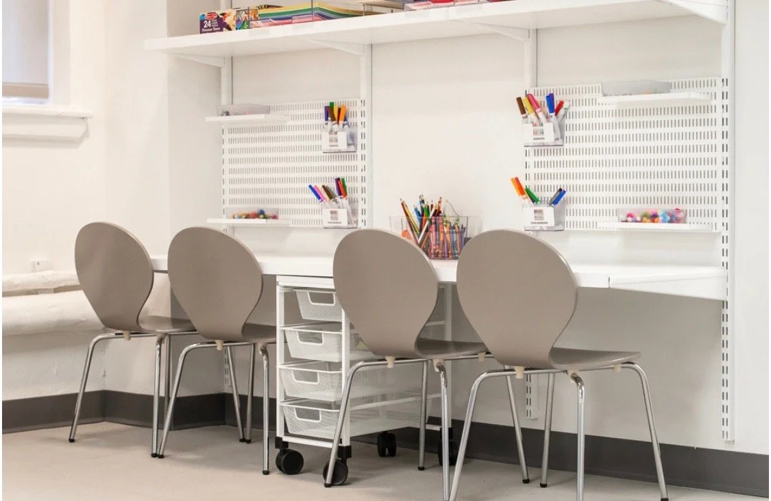 Empty study or work station with four beige chairs, a white desk with drawers, and wall-mounted white pegboards holding colorful markers and supplies in a well-lit room.