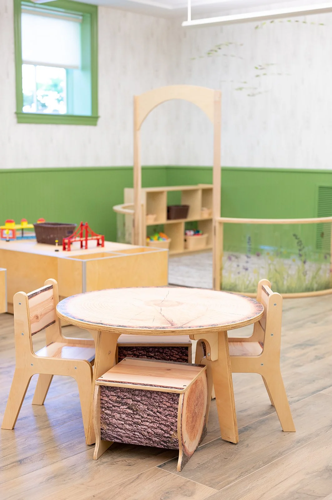 A children's playroom with a round wooden table with log print, surrounded by small wooden chairs, a wooden shelving unit, and toys including a LEGO set and a red bridge, in a room with green and white walls and a window.