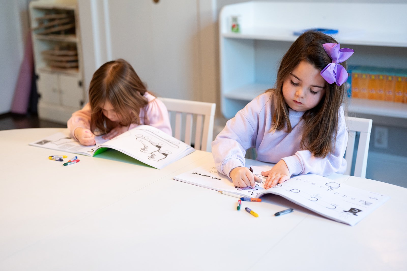 Two young girls sitting at a white table, coloring in their workbooks with crayons, in a classroom setting.