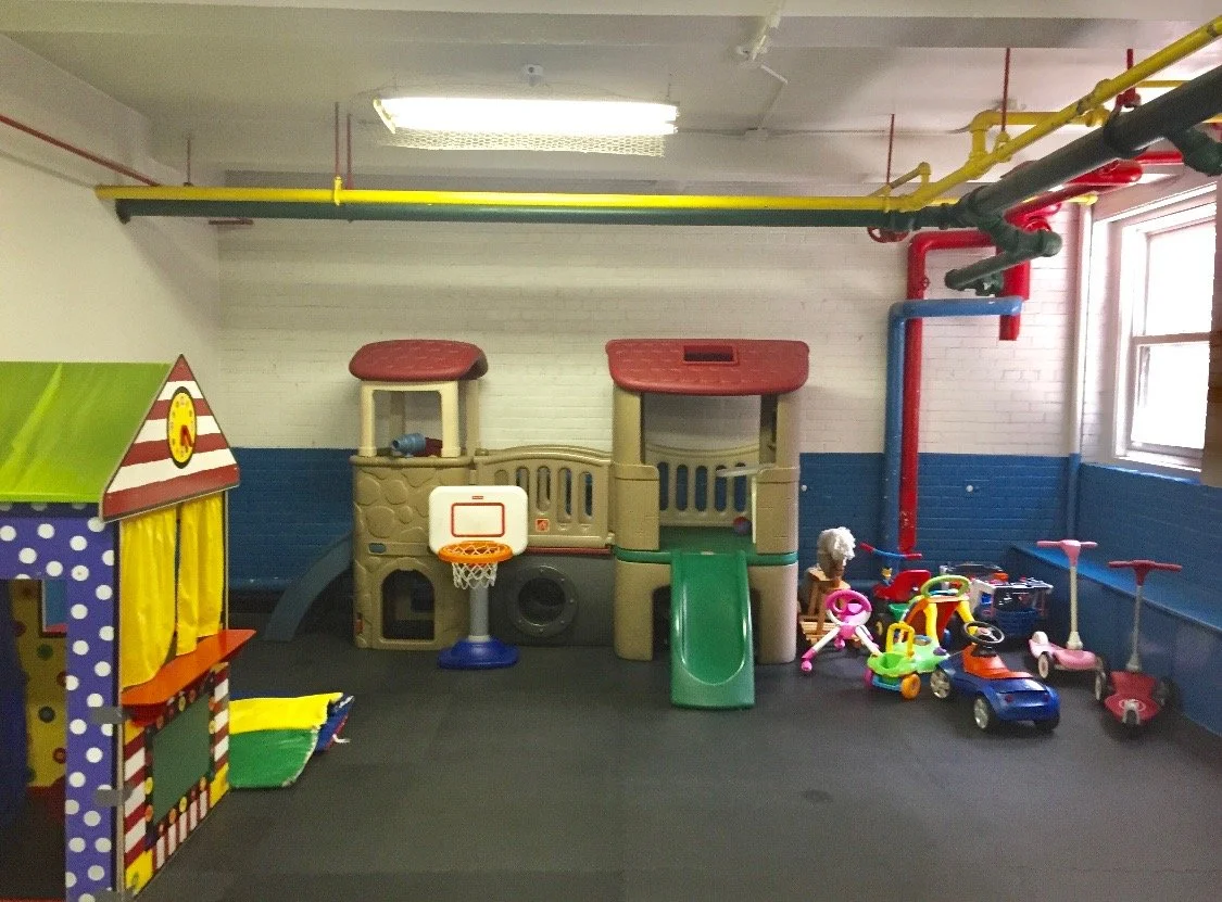 Indoor play area for children with a small plastic slide, a sandbox, and several toy ride-on cars and scooters. There is a colorful playhouse on the left and a window on the right letting in natural light.