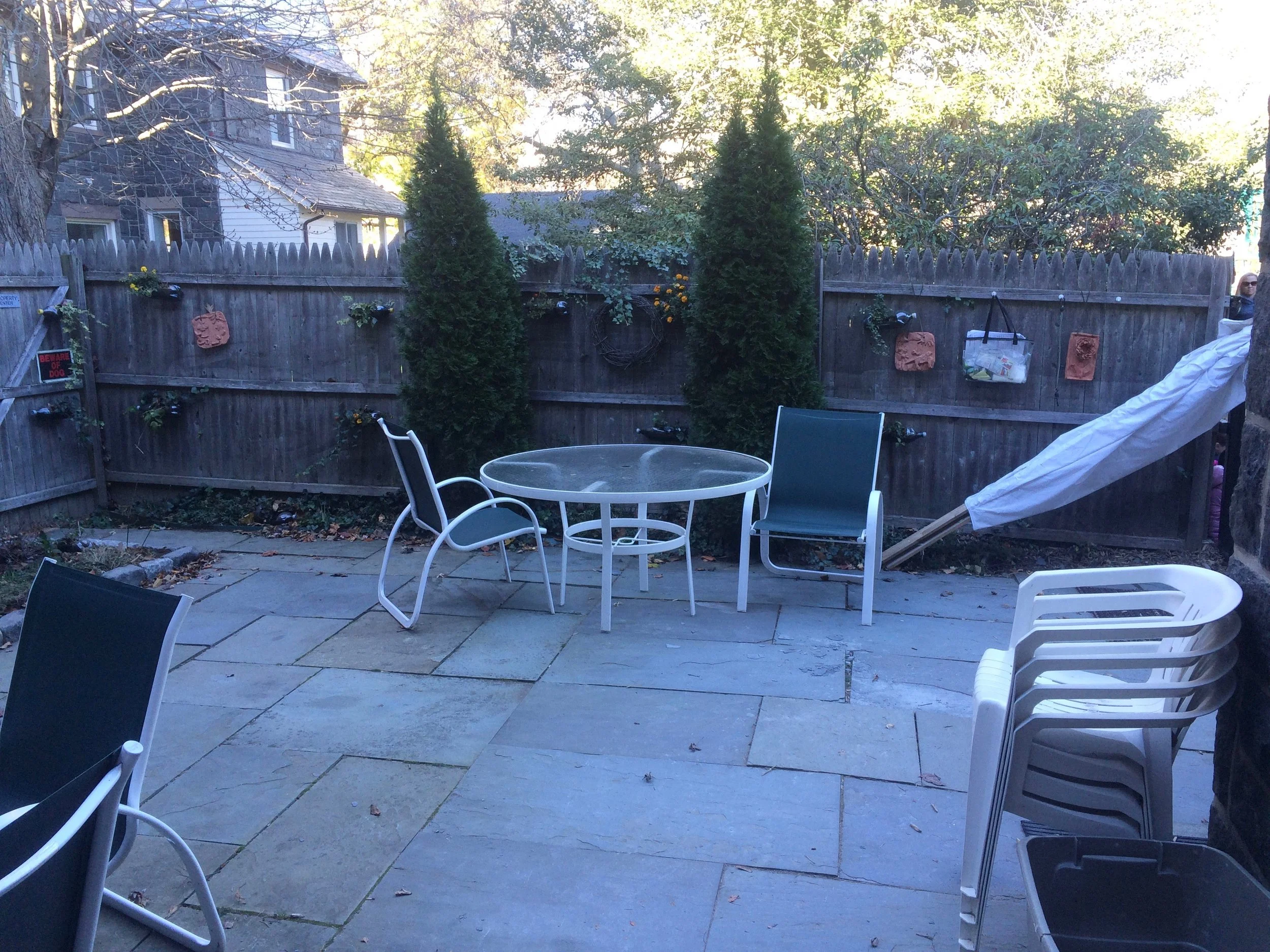 Backyard patio with stacked white plastic chairs, a round glass table, two green metal chairs, and a wooden fence decorated with hanging planters and bricks. Two tall trees and other trees with fall foliage are in the background.