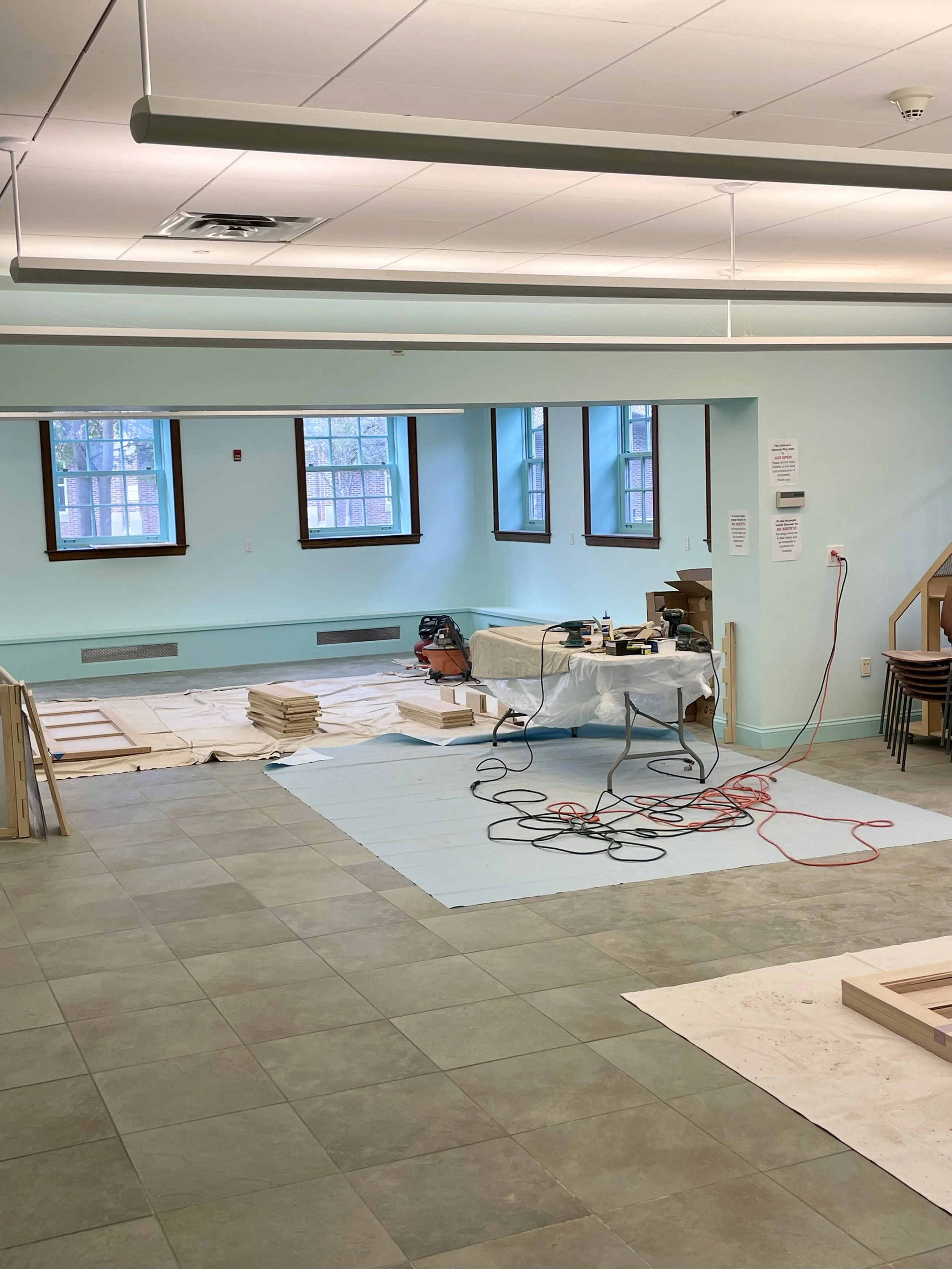 A room under renovation with light green walls, multiple windows, construction tools, and materials on the floor and table, with some chairs stacked near the staircase.