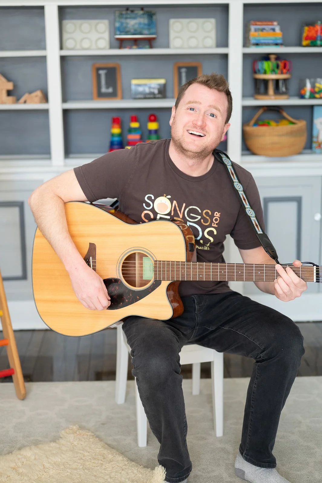 A man smiling while sitting on a white chair and playing an acoustic guitar in a cozy room with a bookshelf in the background.