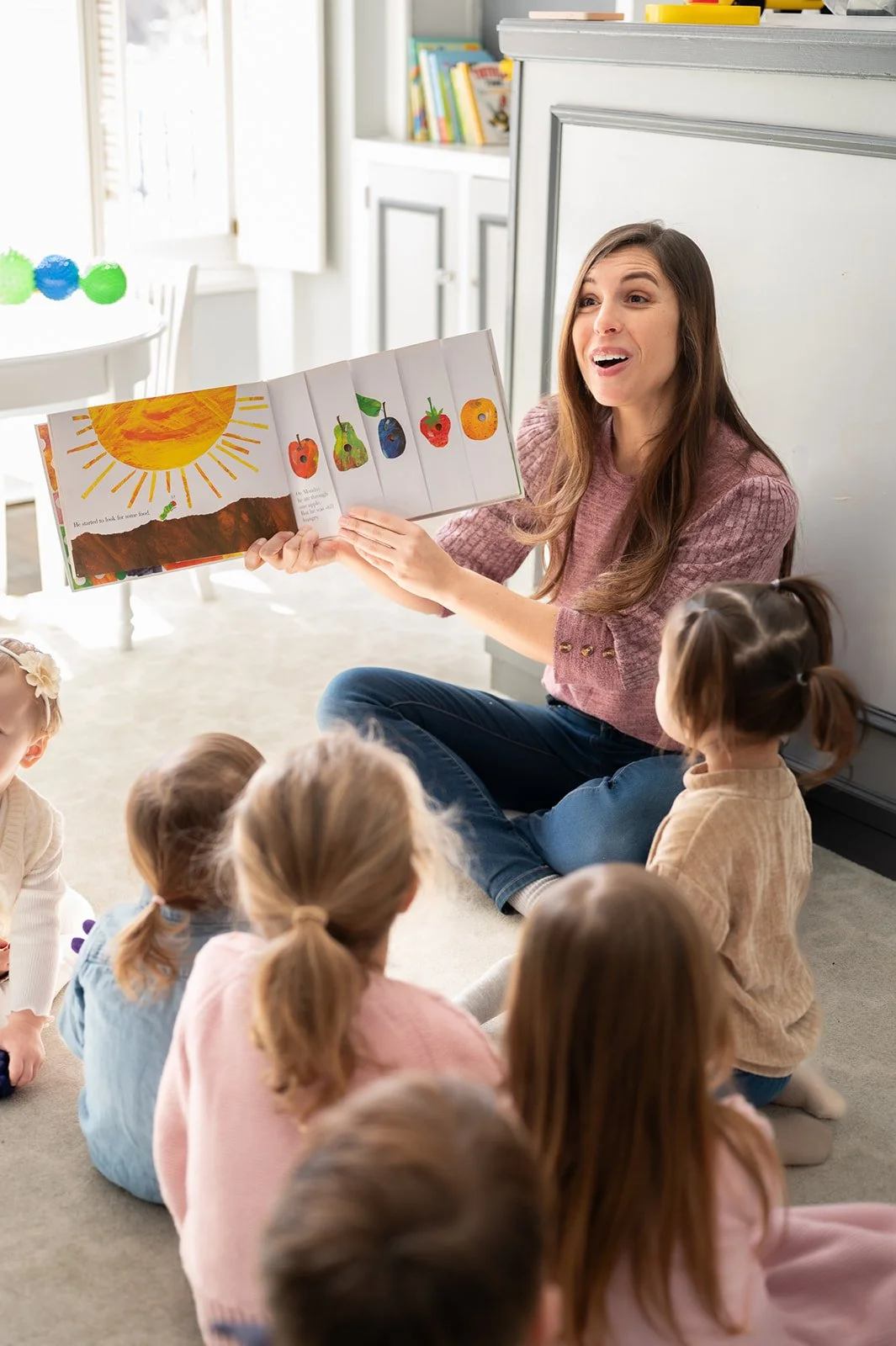 A woman reading a picture book to a group of children sitting on the floor in a bright room.