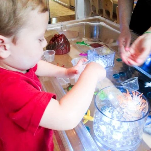 Child playing with colorful plastic toys and slime at a clear plastic table.