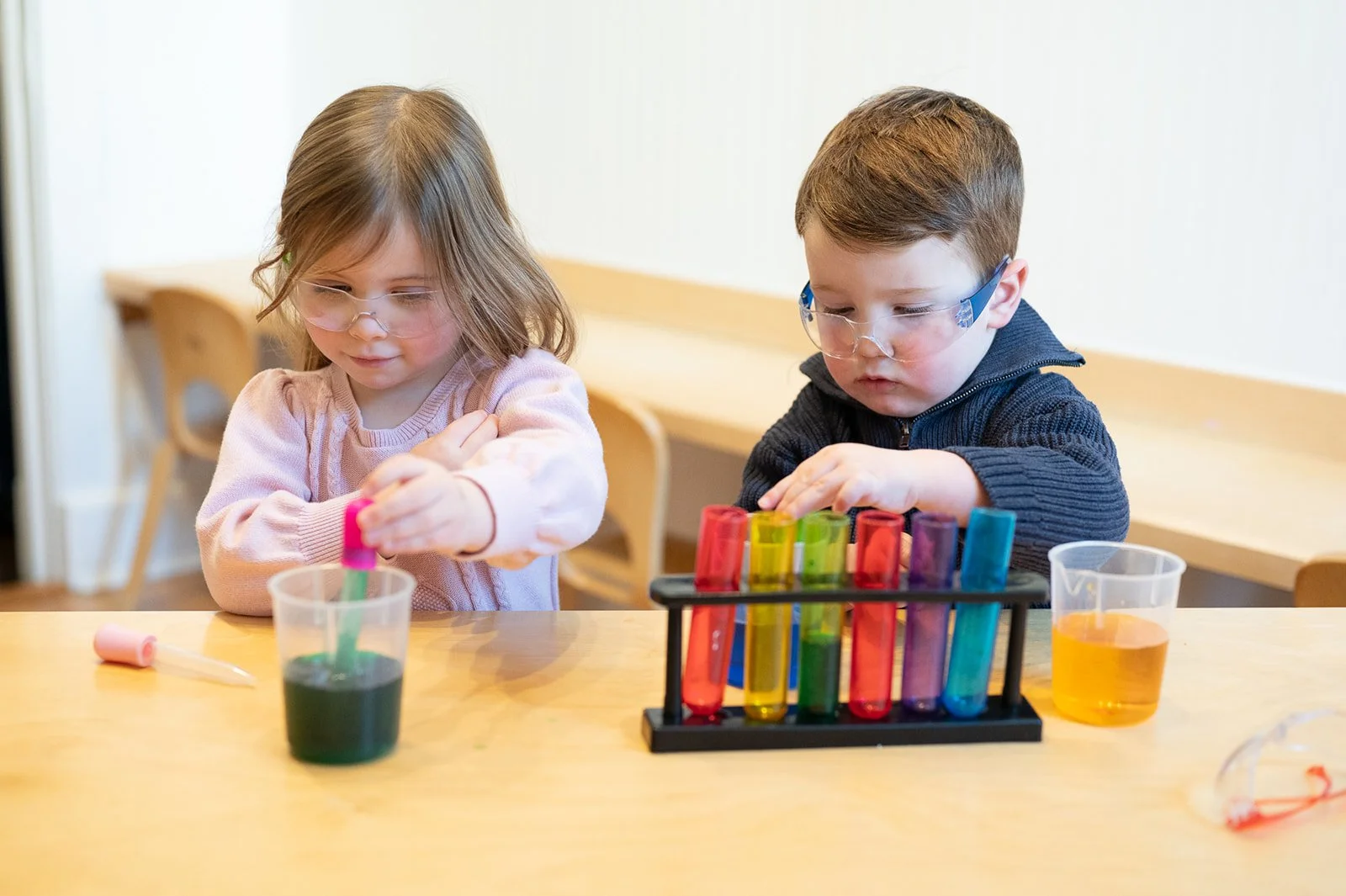 Two young children wearing safety glasses are conducting a science experiment with colorful liquids and test tubes.