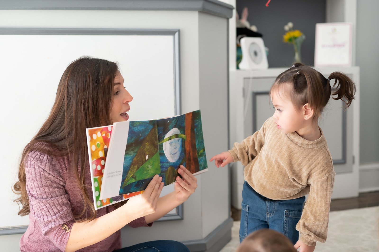 A woman holding a colorful children's book with a landscape illustration, engaging with a young girl pointing at the book, in a cozy room.