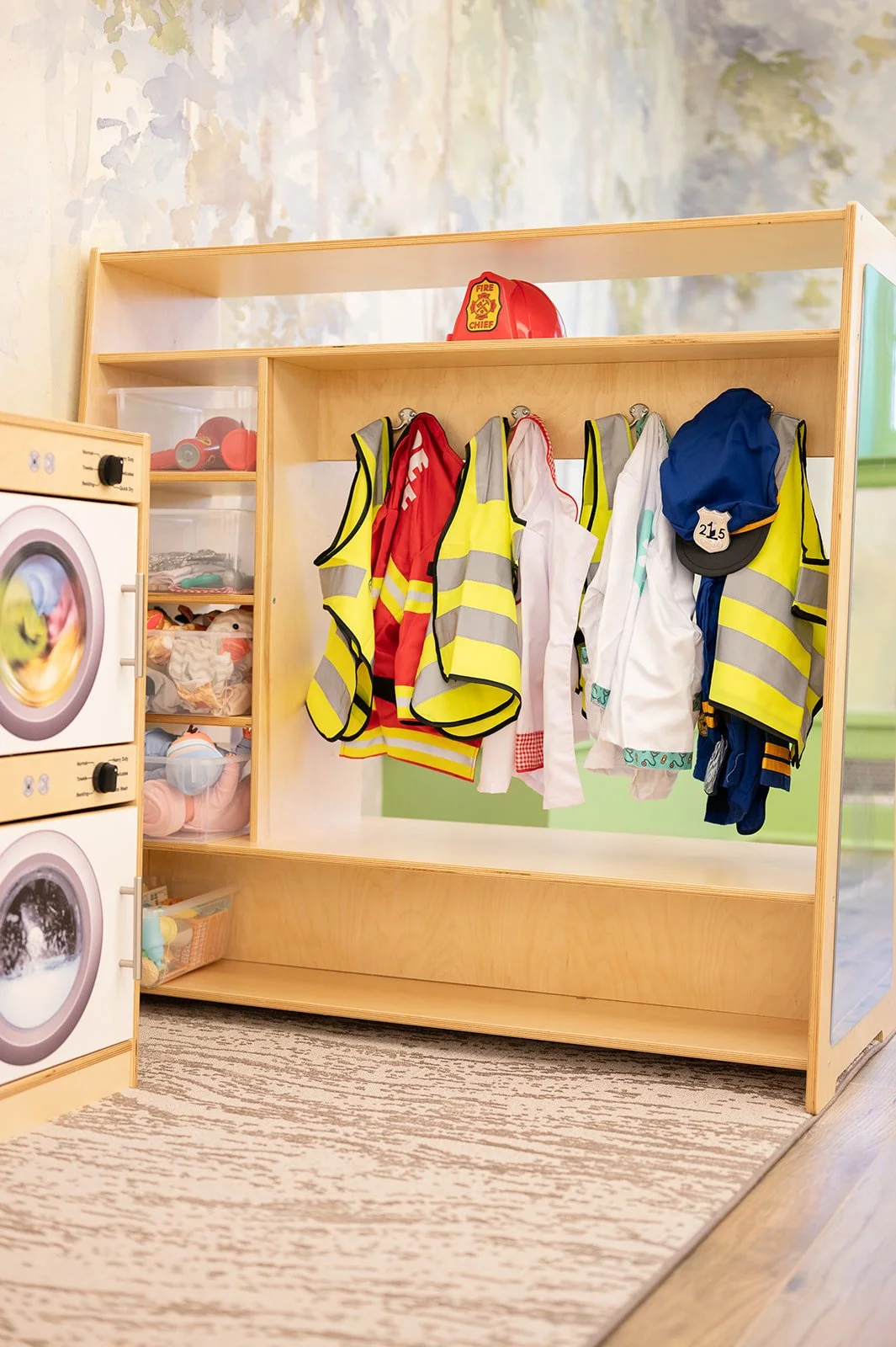 Children's dress-up corner with firefighter costumes, safety vests, and helmets on a wooden rack, next to a toy washing machine and shelves with toys, in a classroom or playroom.