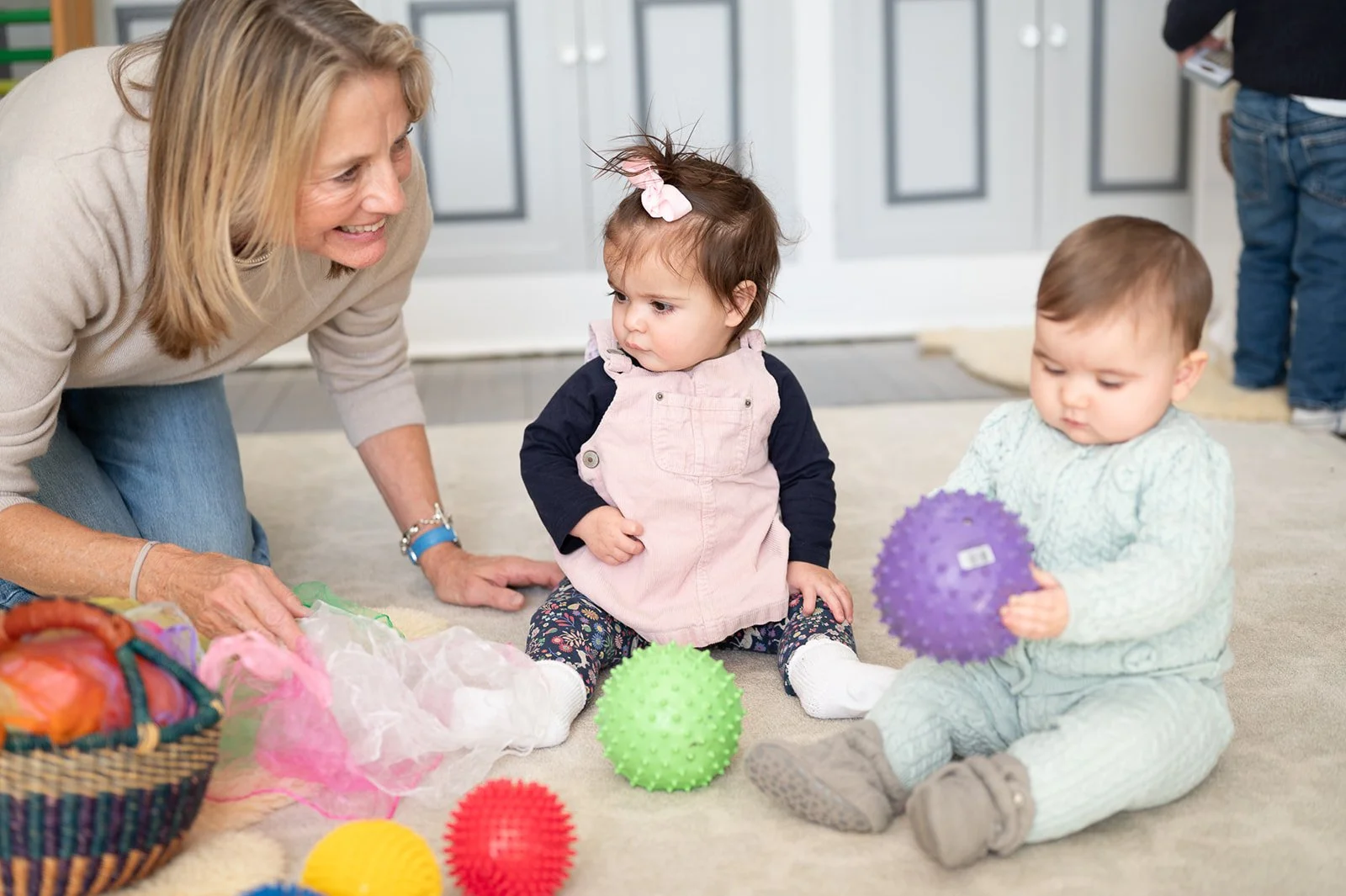 An elderly woman playing with two young children on the floor, surrounded by colorful textured balls and a basket of yarn, in a cozy home setting.