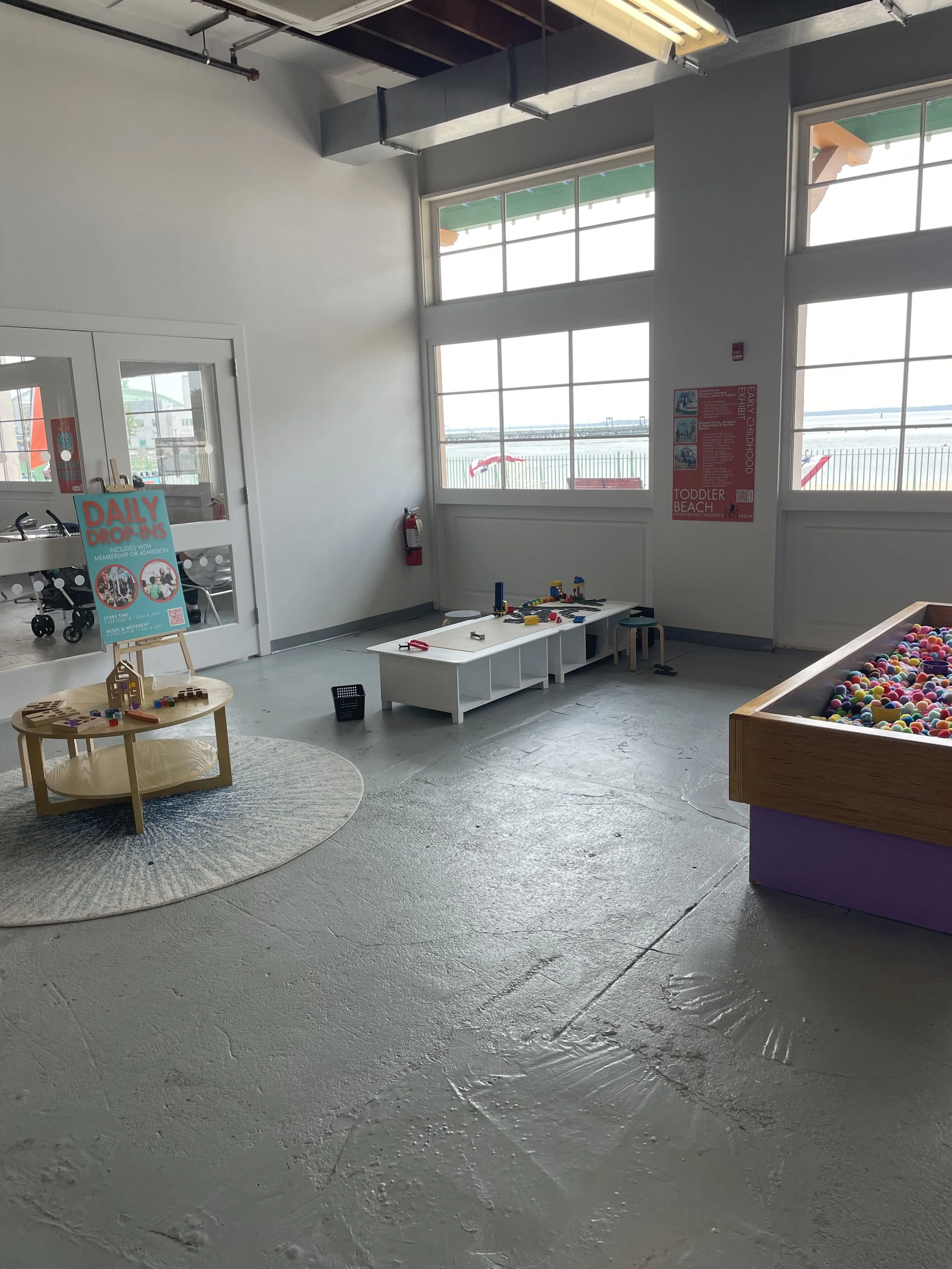 Indoor children's play area with a small white table, a ball pit, a play mat, and a sign advertising daily drop-ins, near large windows overlooking a body of water.
