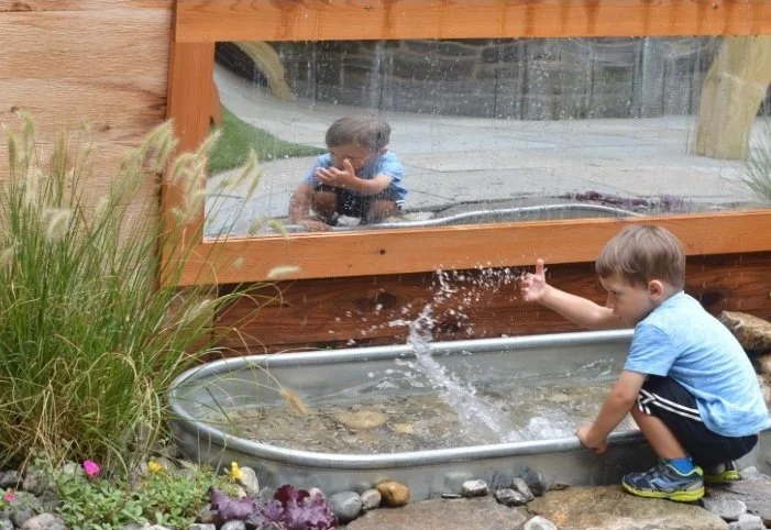 A young boy playing with water in a small outdoor pond near a wooden and glass structure that reflects his image.