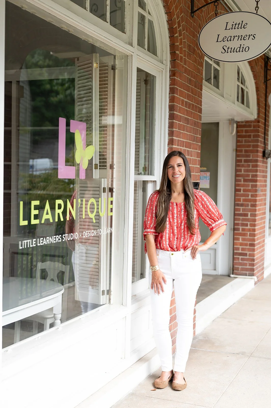 A woman standing outside a storefront with a sign that reads "Little Learners Studio". The woman is smiling, wearing a red patterned blouse and white pants, and standing on a sidewalk in front of a brick building with large windows.