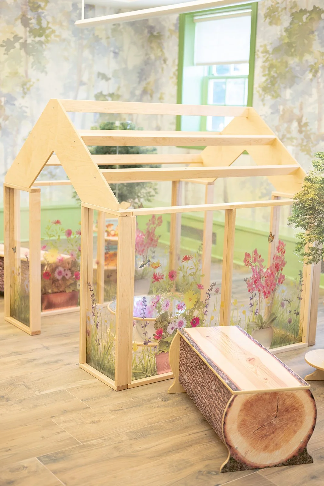 Small wooden house frame with transparent sides decorated with floral patterns, sitting inside a room with a window and wooden floor, and a log-shaped bench in front.